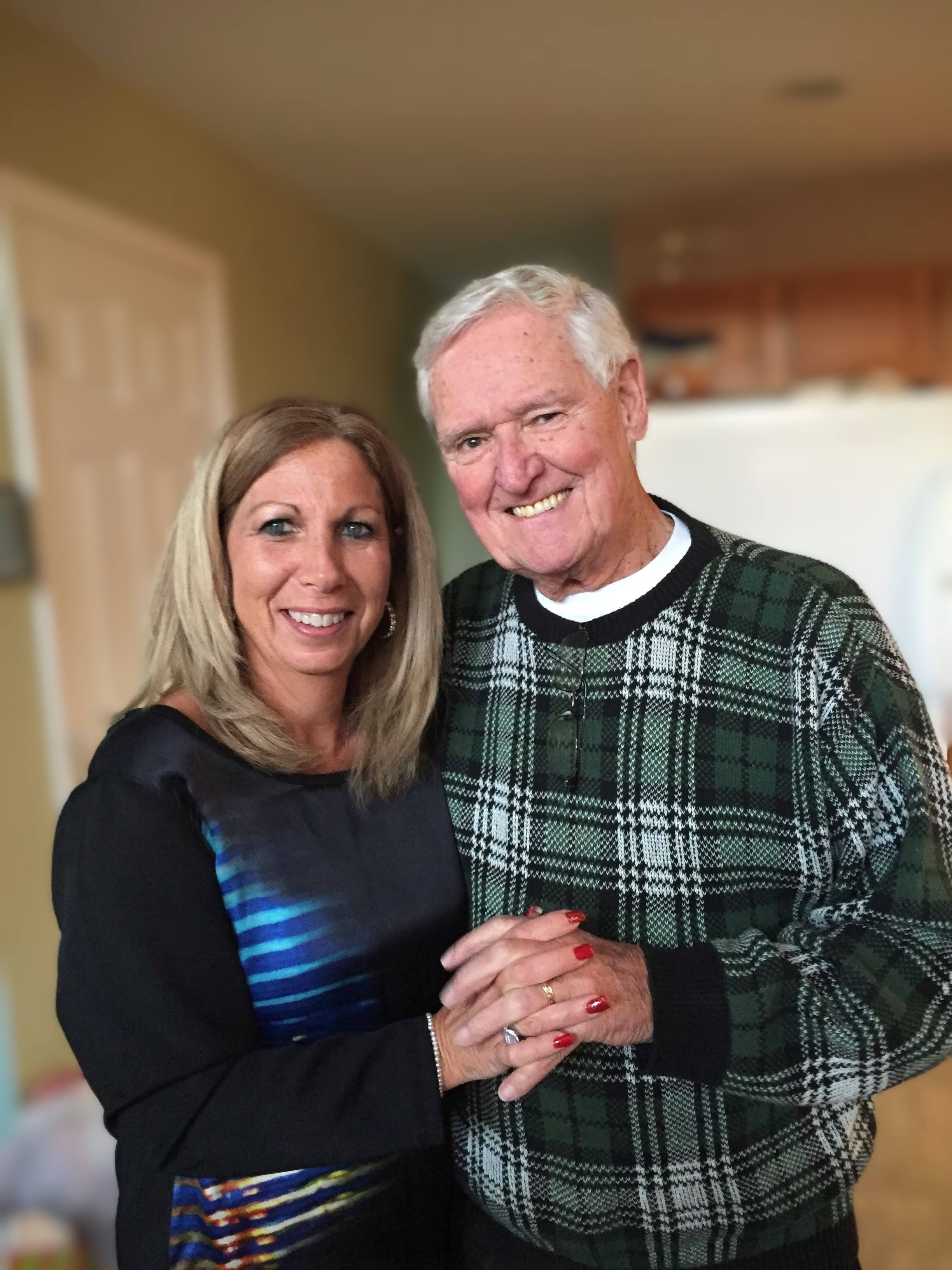 A smiling couple, a woman with blonde hair and a man with white hair, holding hands and standing close in a kitchen setting.