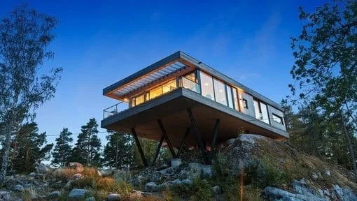 Modern house built on stilts on a rocky hill, surrounded by trees, under a clear blue sky.