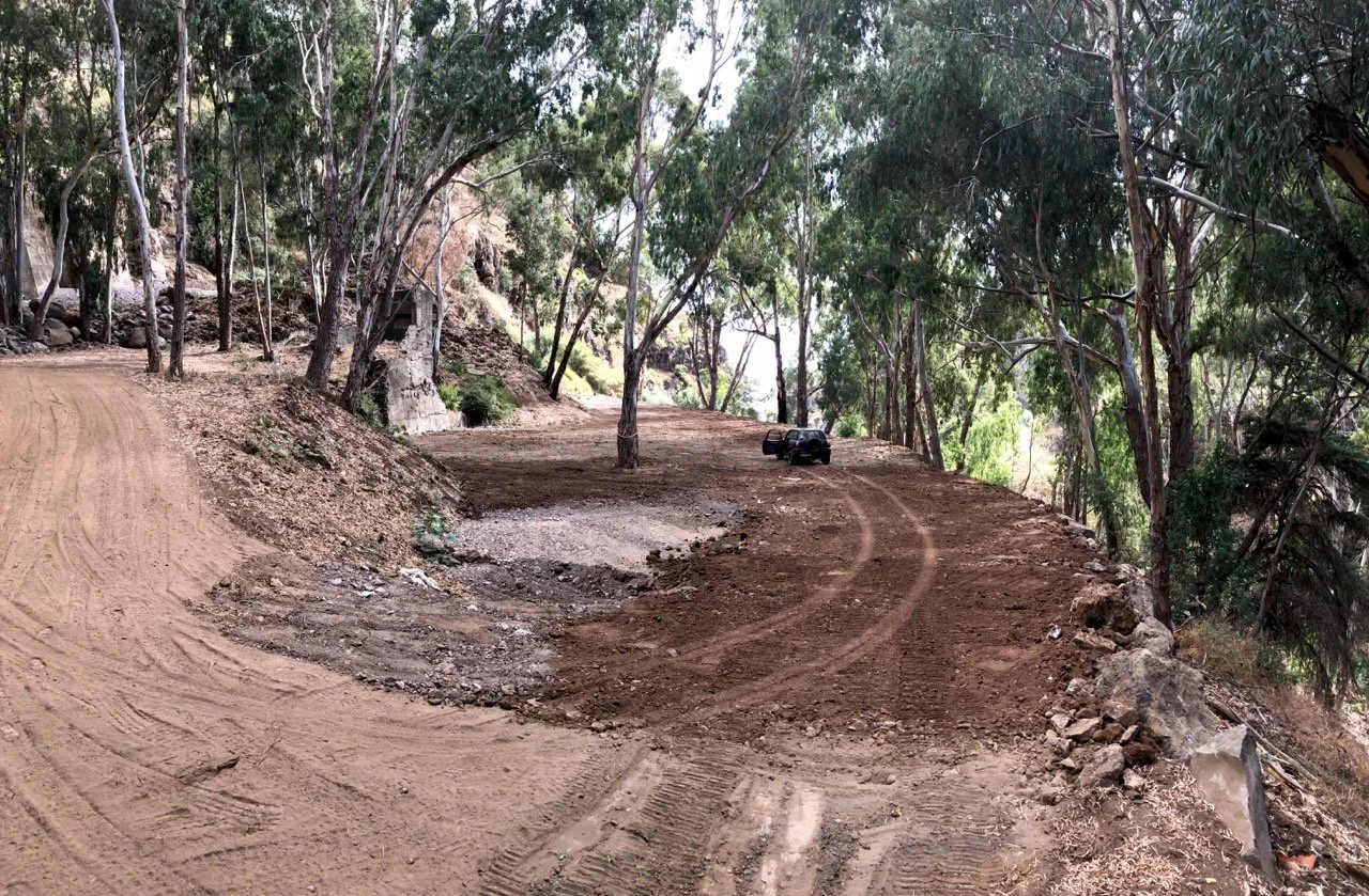 A winding dirt road through a wooded area with trees and rocks, and a black vehicle parked on the side.