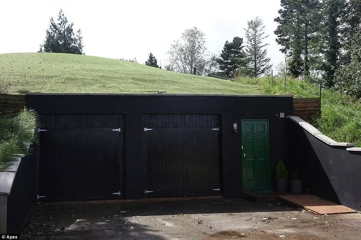 A black garage door and a green door on a modern house built into a hillside with a grassy roof and surrounded by trees.