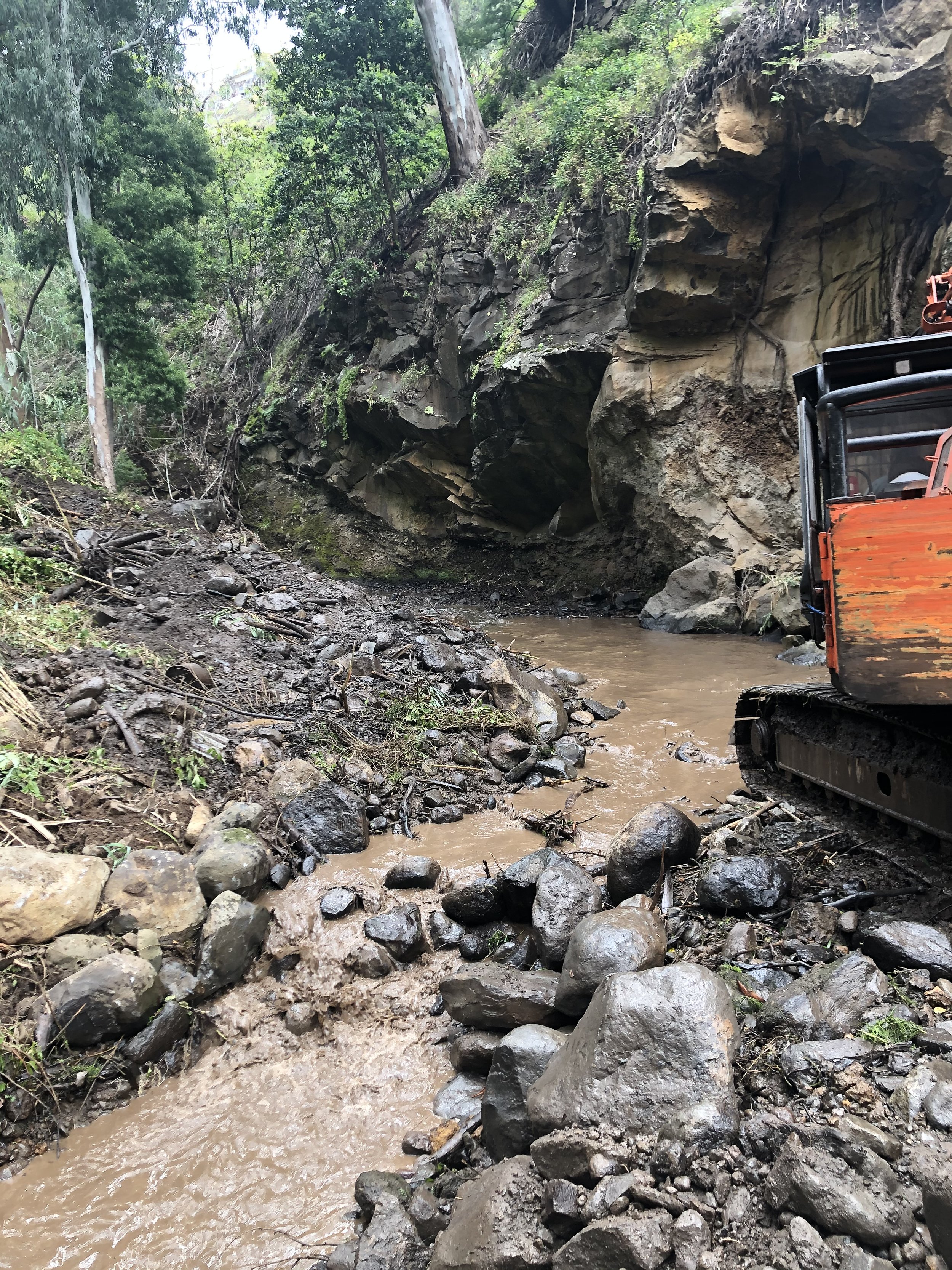 A muddy construction site beside a rocky hillside with a small water stream, a bulldozer partially visible on the right, and lush green trees in the background.