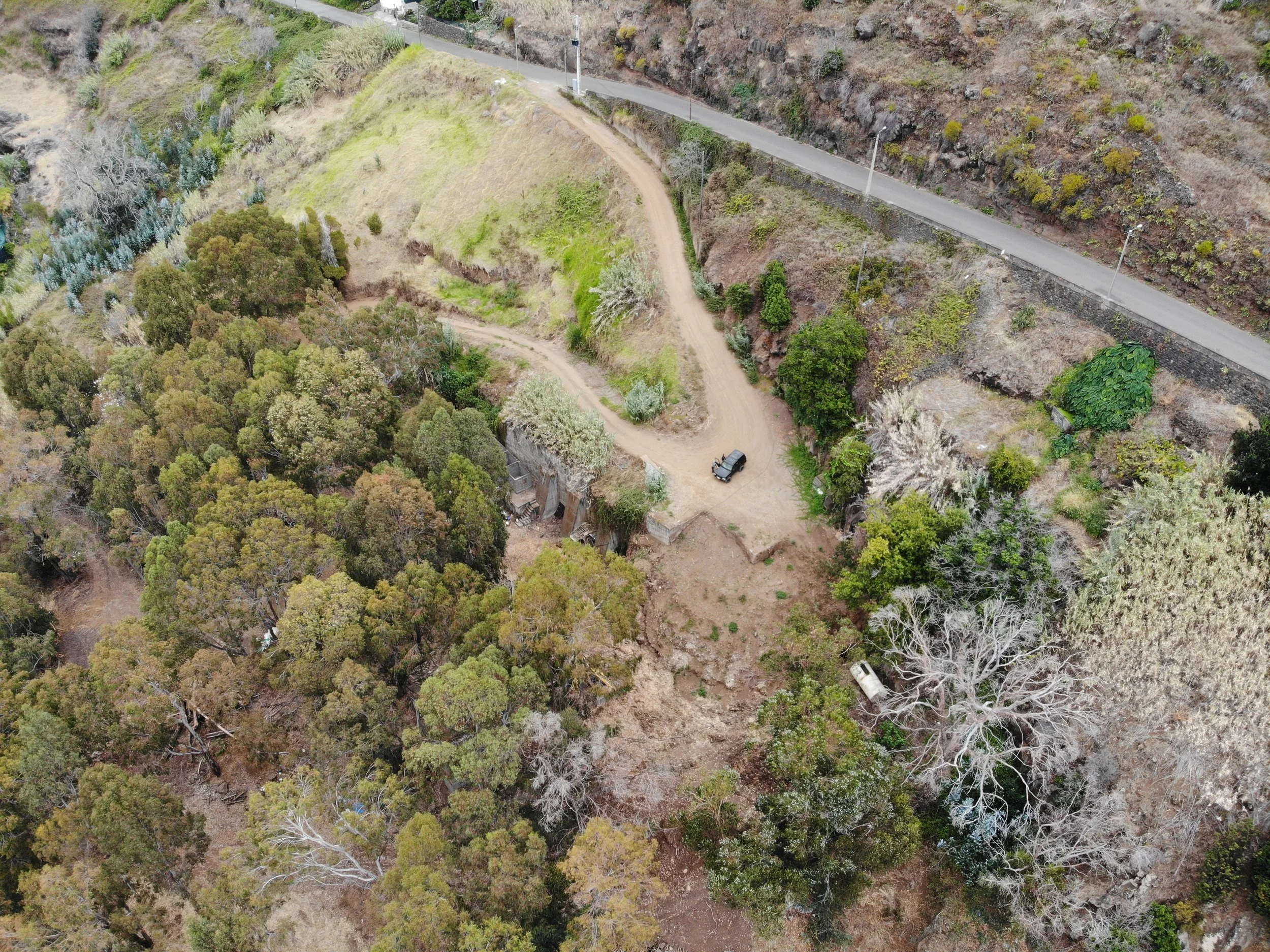 Aerial view of a rugged landscape with dirt paths, a parked vehicle, dense trees, and a paved road on the hillside.