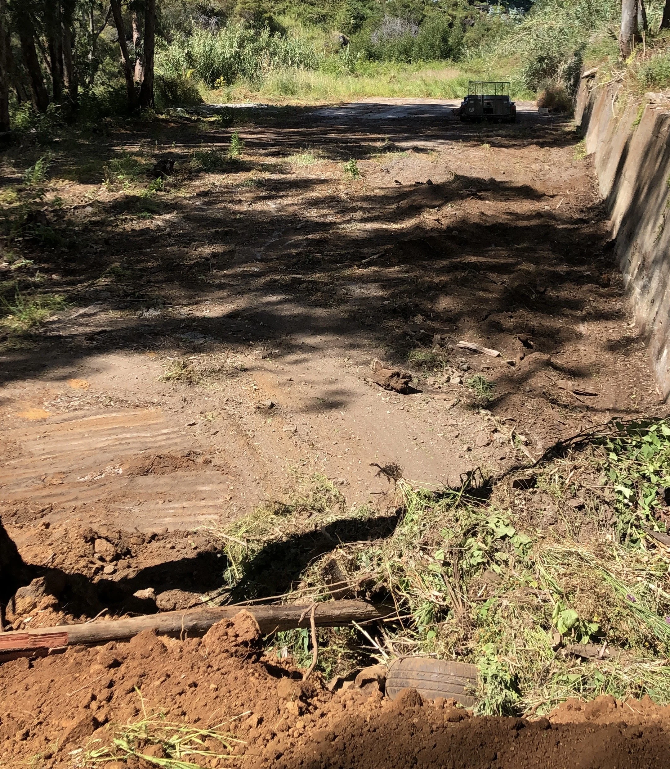 A dirt pathway in a shaded outdoor area, with a small trailer in the distance and a broken tire at the bottom foreground, surrounded by trees and greenery.