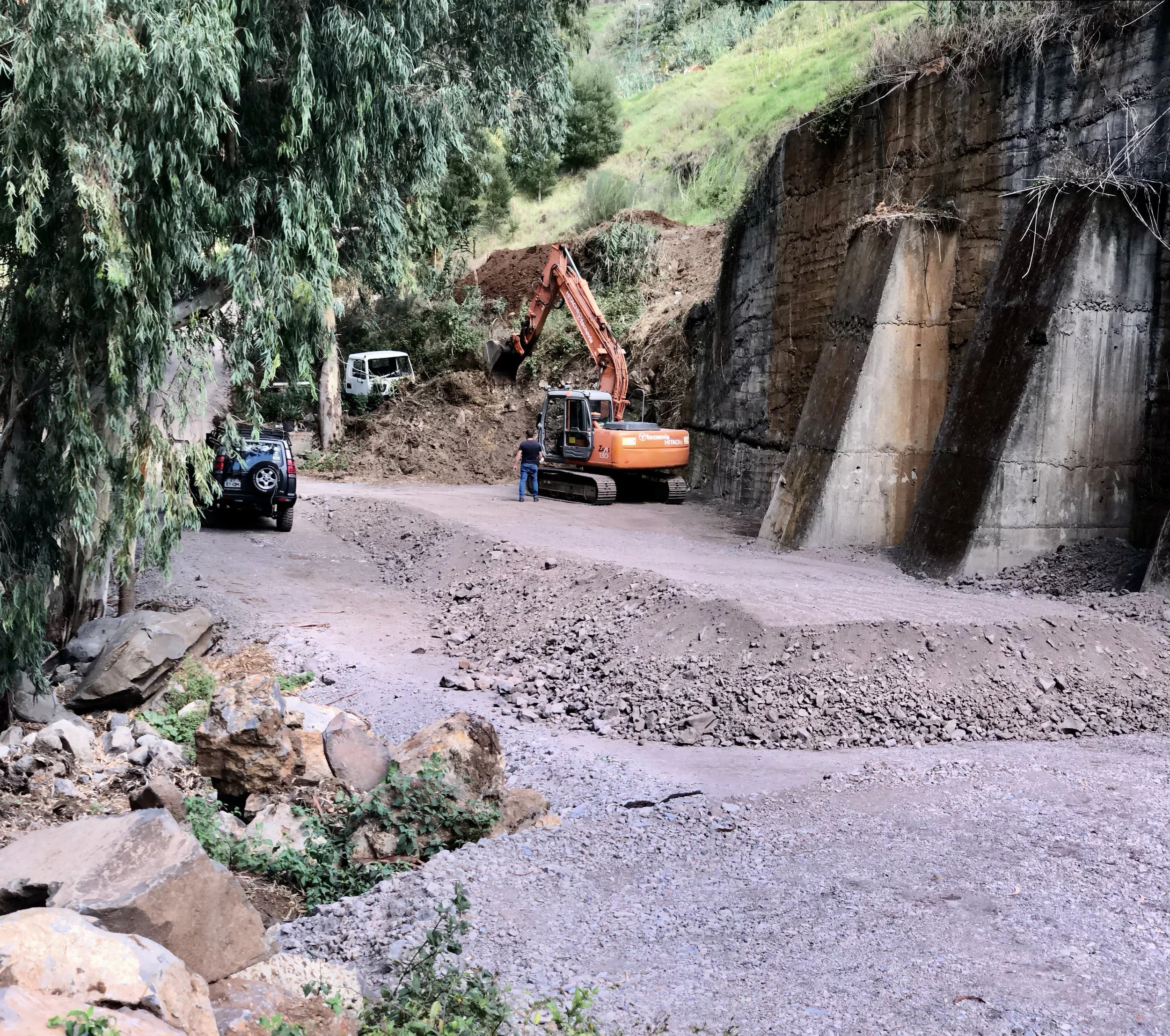 Construction workers and excavator working on a dirt road next to a large rock face, with parked vehicles nearby.