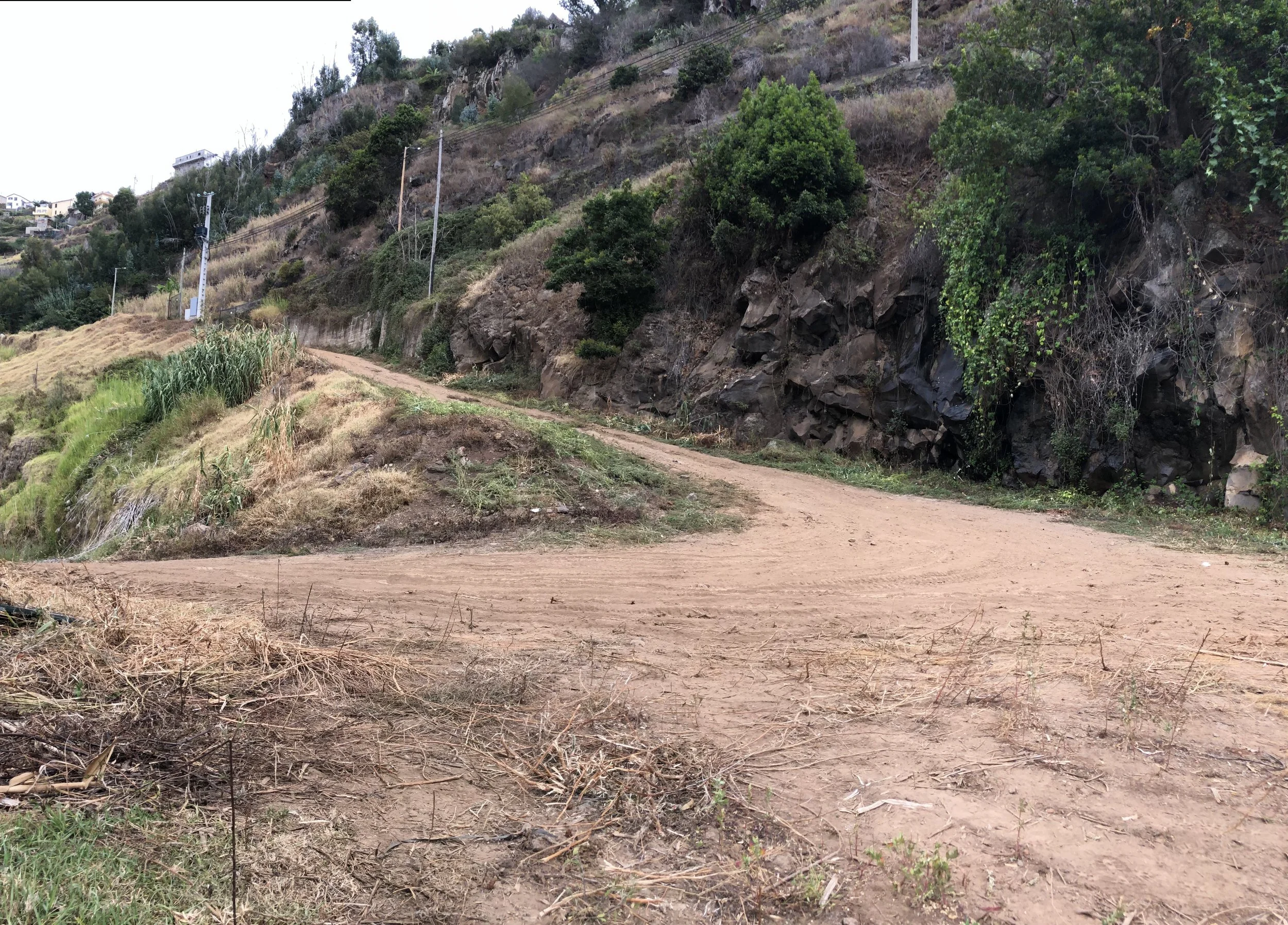 A dirt road winding along the side of a hill with rocky and green vegetation on the hillside. There are utility poles with power lines running parallel to the road. In the background, there are residential houses on the hilltop.