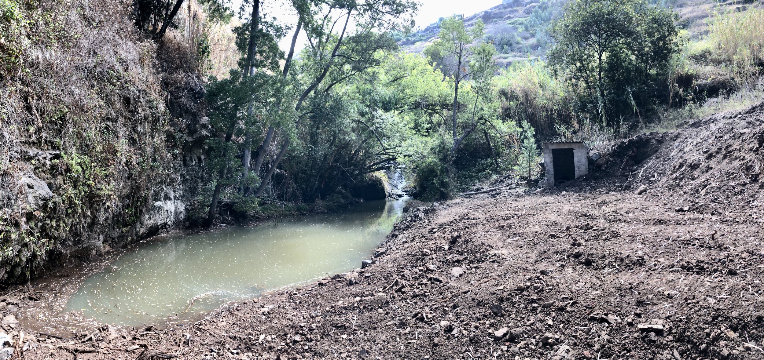 A small waterway flowing through a rocky landscape with trees and shrubs on both sides, and a small concrete structure near the bank.