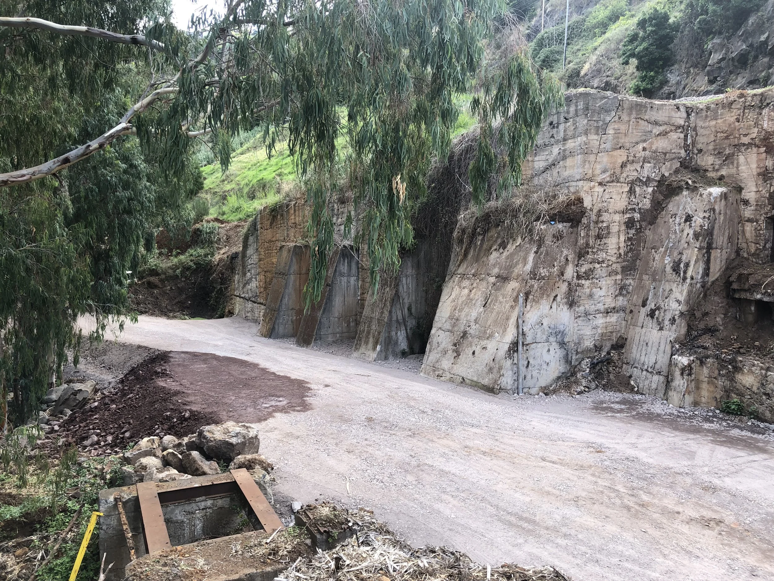 A dirt road runs past a rocky hillside with large concrete retaining walls, surrounded by green trees and vegetation.