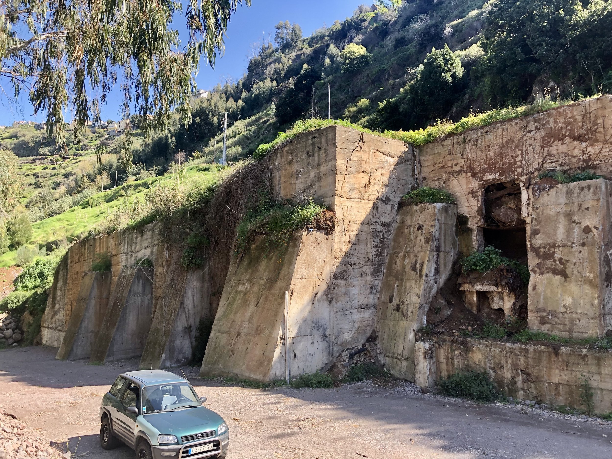 A small gray car parked on dirt near a large, old concrete structure with multiple openings, set against a hillside with lush green vegetation and trees in the background.