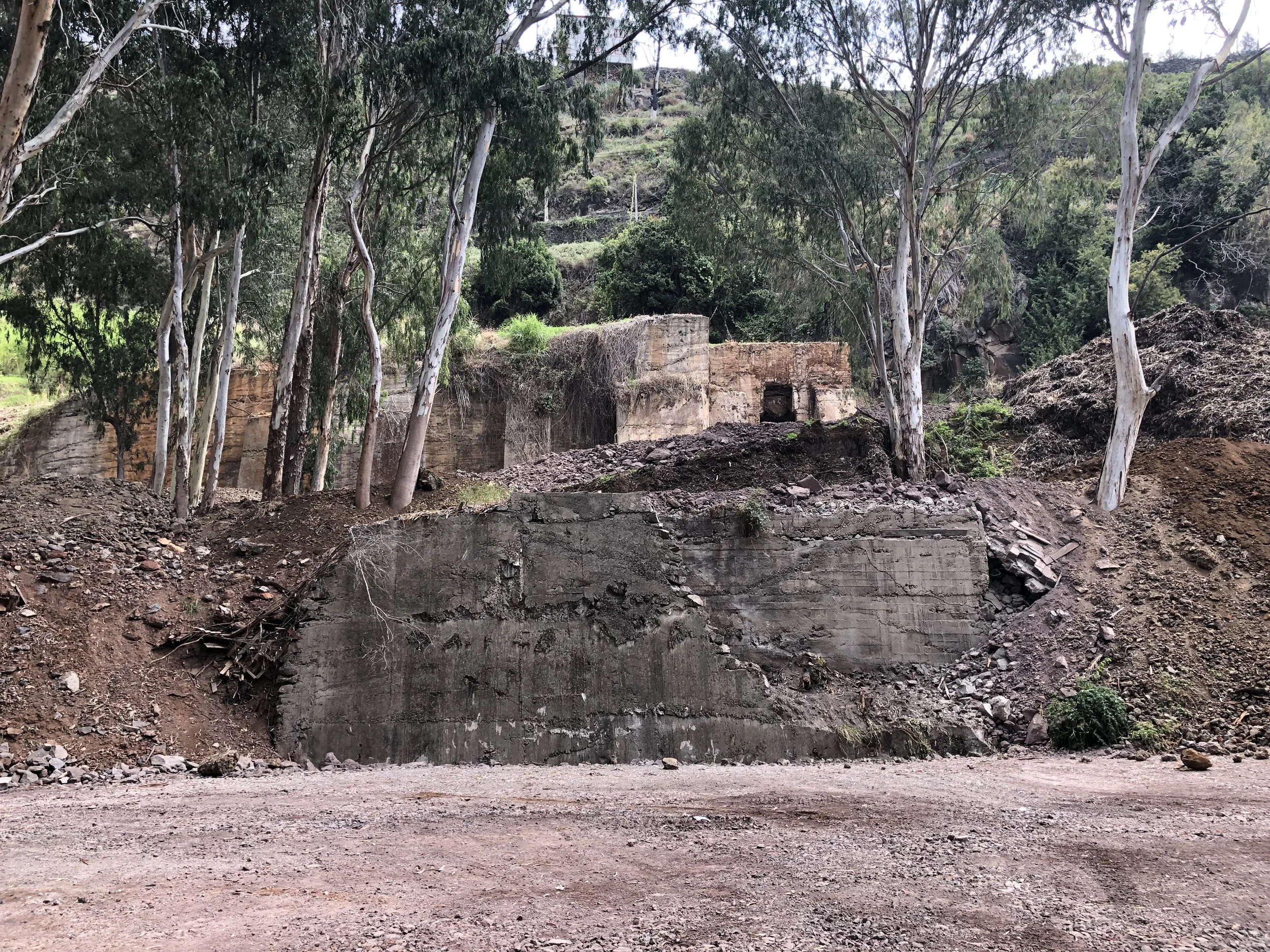 Ruined concrete structure on a hillside with trees and shrubs around, and a dirt road in the foreground.