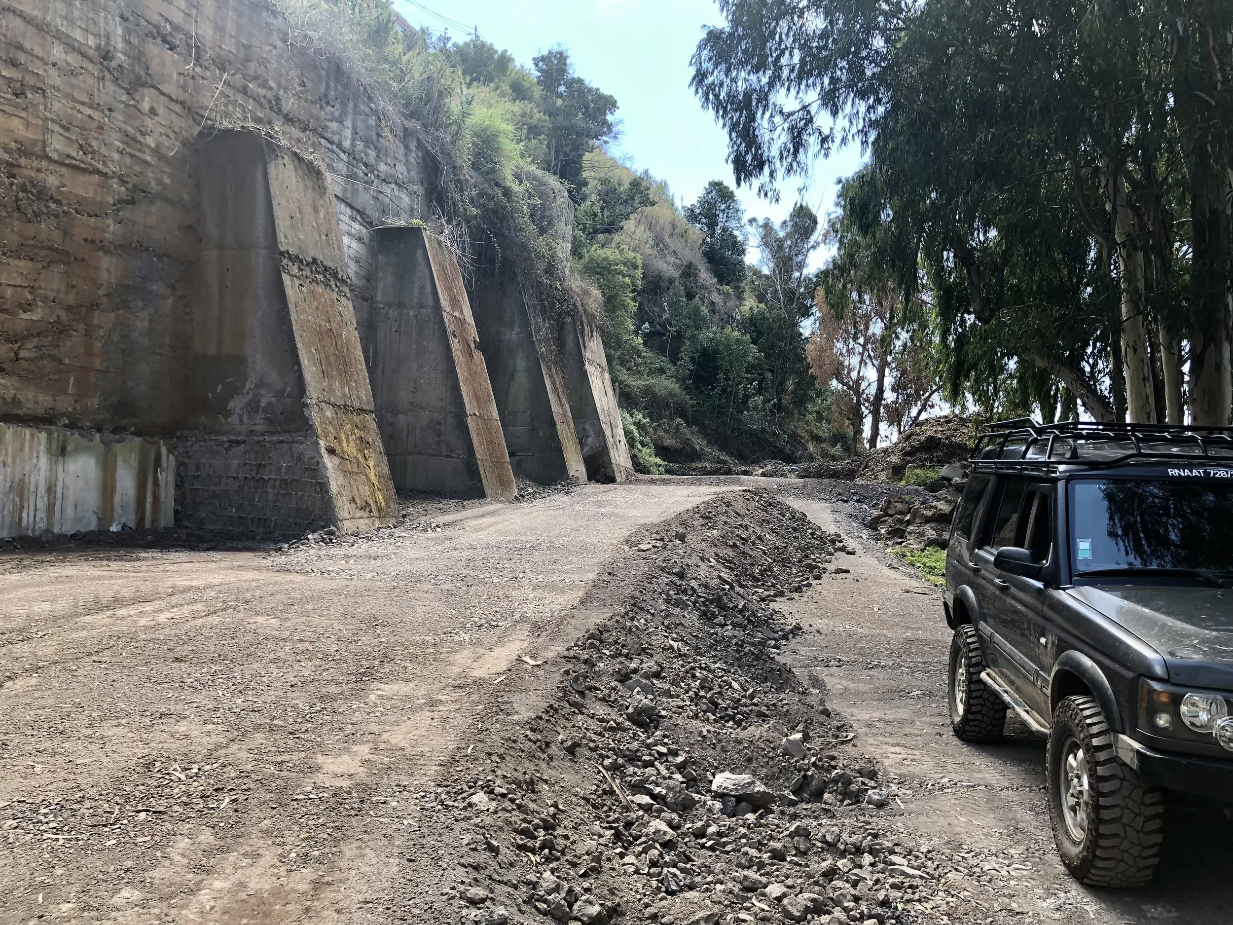 A dirt road with a pile of rocks in the middle, a black SUV on the right, tall concrete supports along the left side, trees on the right, and a hillside with vegetation in the background.