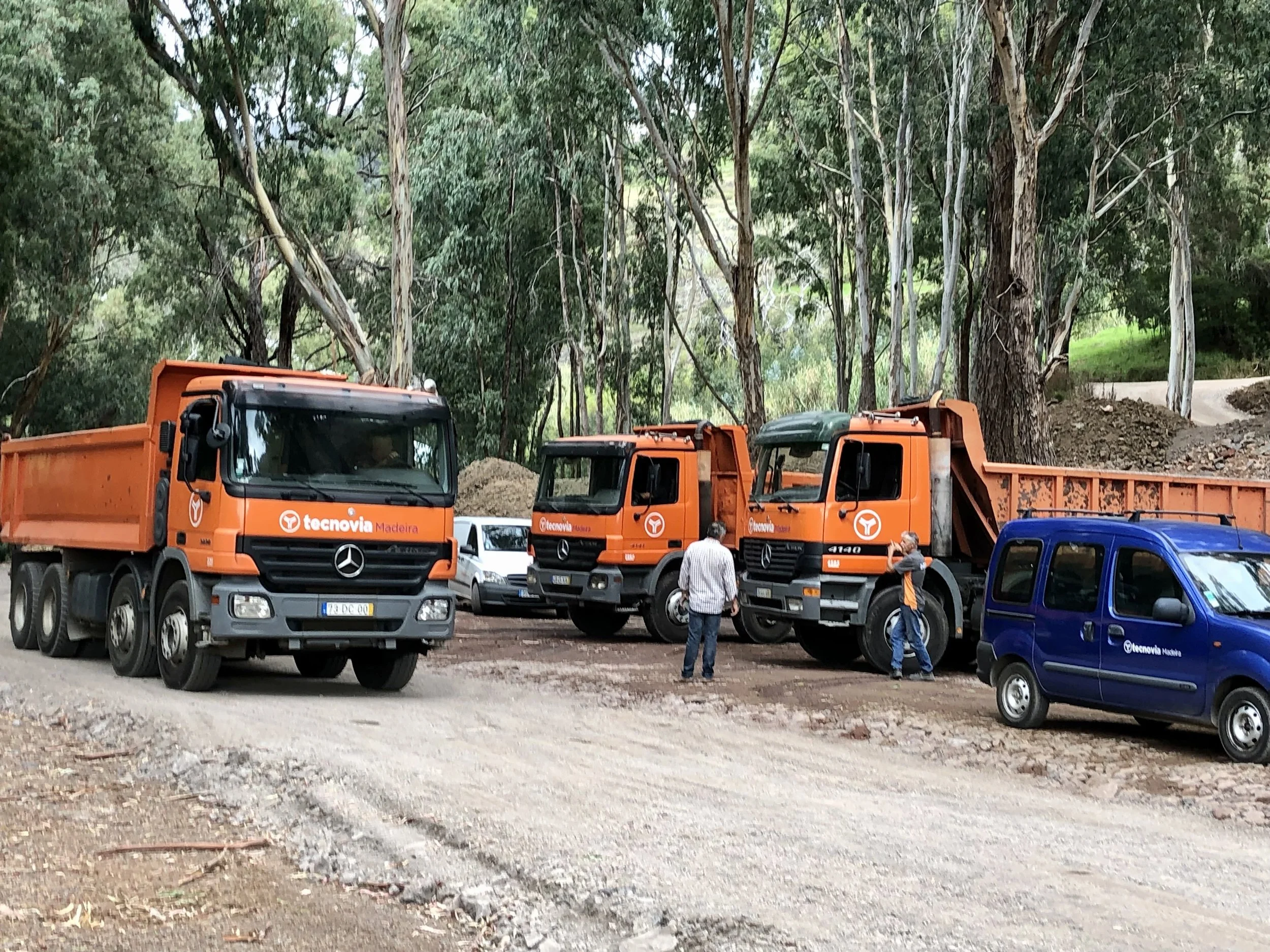 Group of trucks and vans parked on a dirt road in a forested area, with two men standing and talking near the trucks.