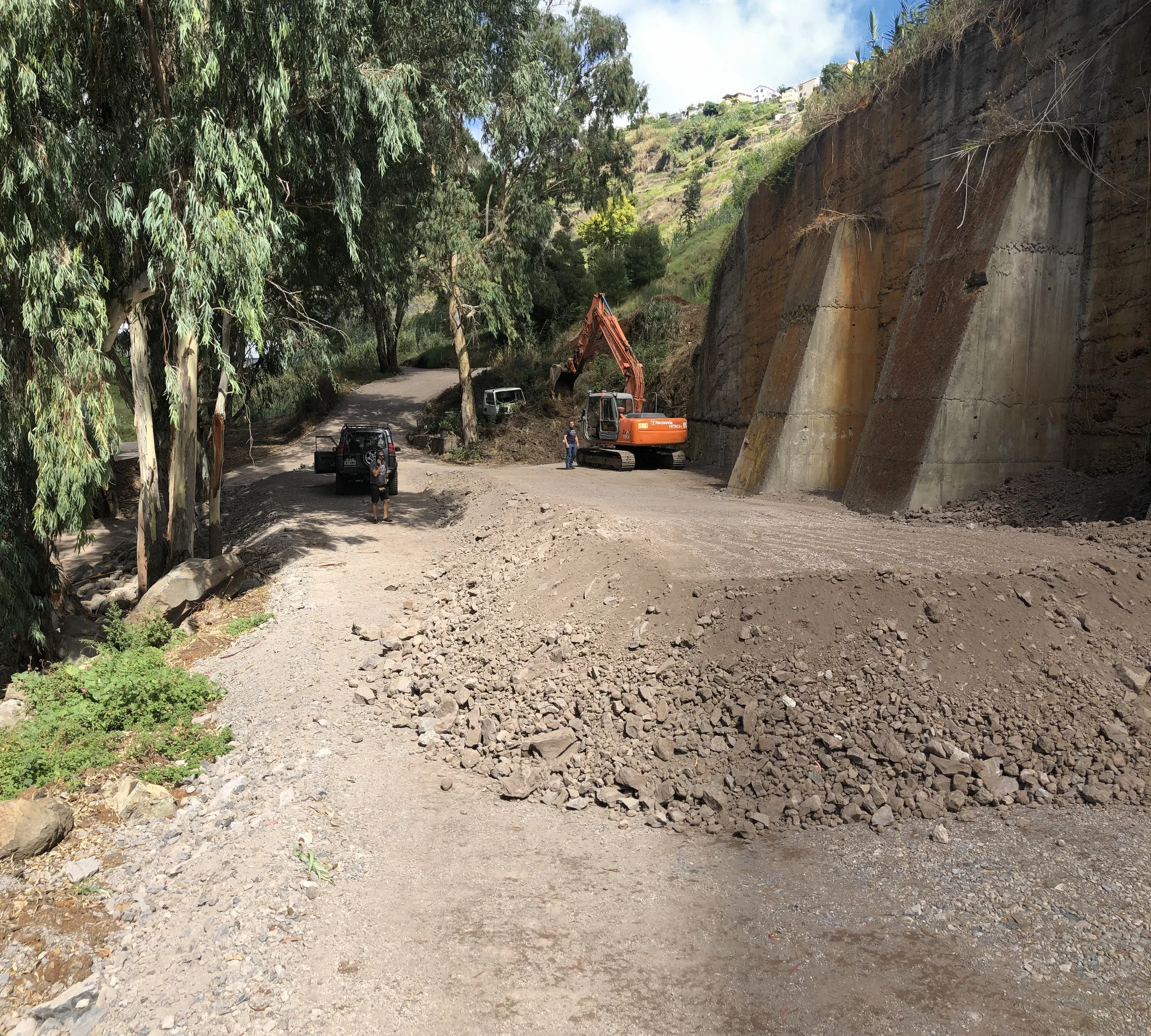 Construction site with excavator working on a dirt road beside a rocky cliff, surrounded by trees and hills in the background.