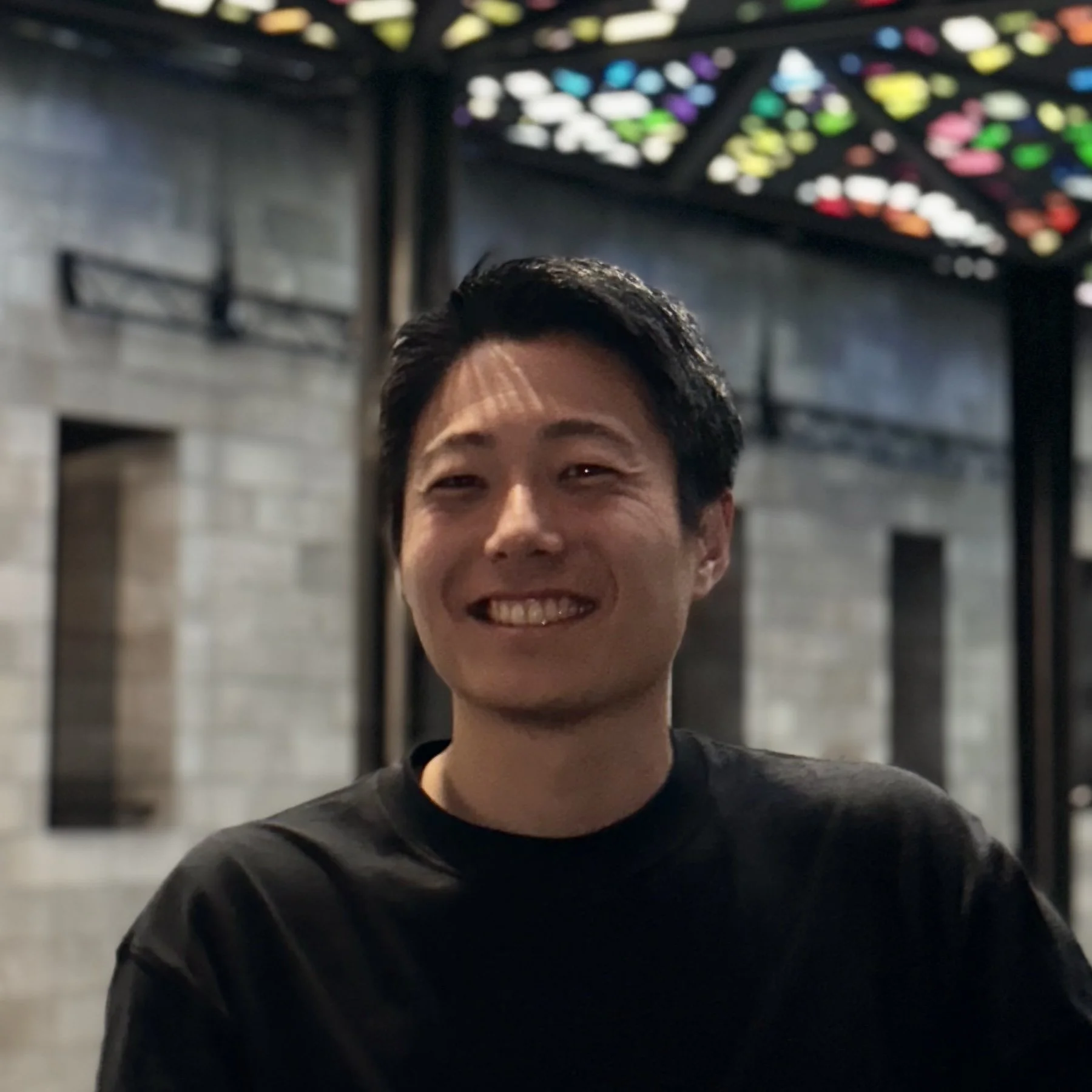 笑顔の若い男性が屋内で写真を撮っている。背景には明るいステンドグラスの天井と石の壁が映っている。