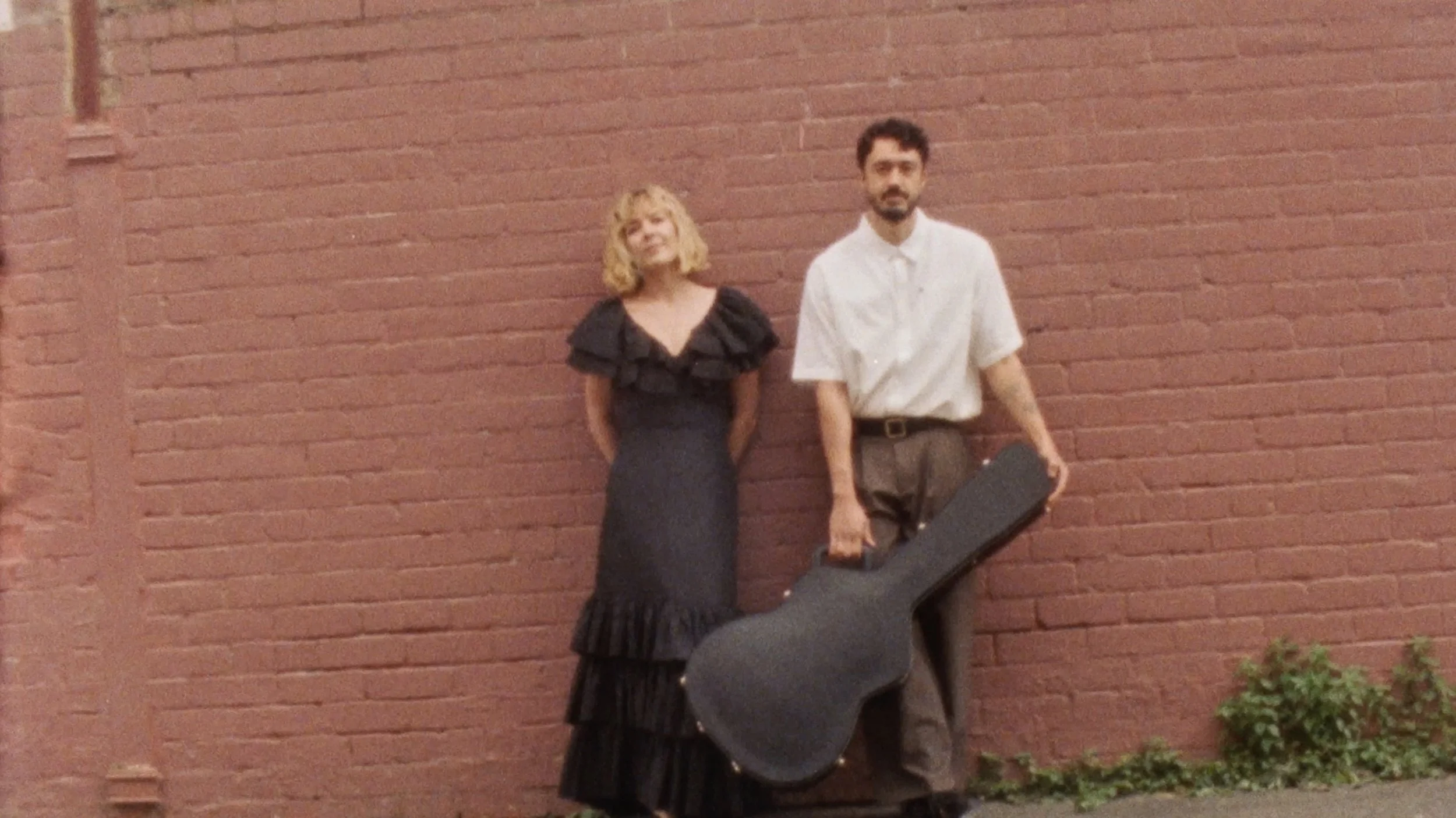 A woman in a black ruffled dress and a man in a white shirt and brown pants standing against a red brick wall. The man is holding a guitar case.