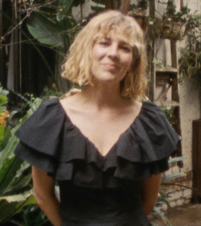 Jess with curly blonde hair smiling outdoors, wearing a black ruffled top, with plants and a wooden fence in the background.