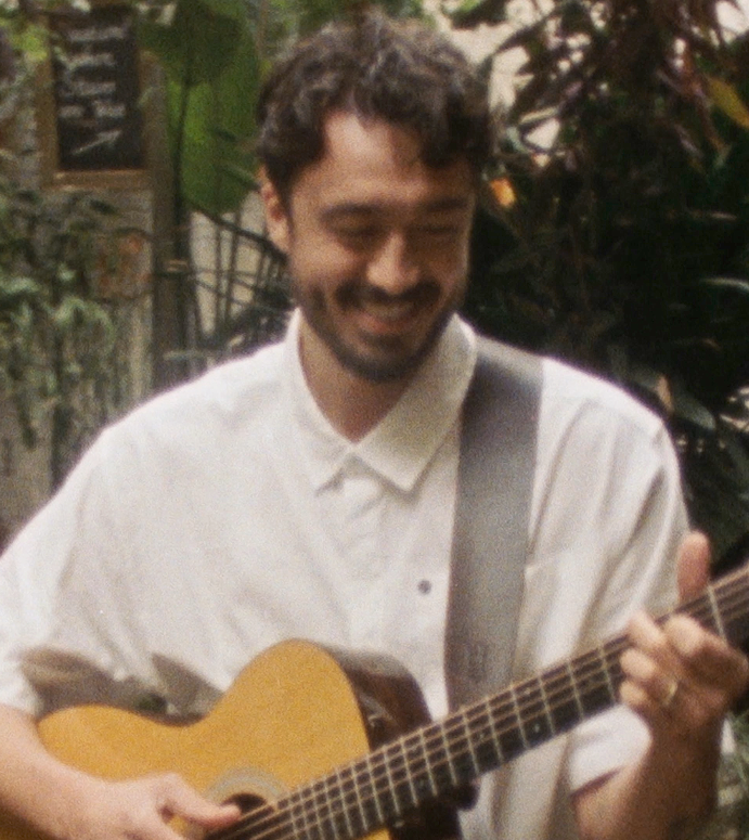 A photo of Nick. He has dark, curly hair and a beard smiling while playing an acoustic guitar outdoors, wearing a light-coloured button-up shirt.