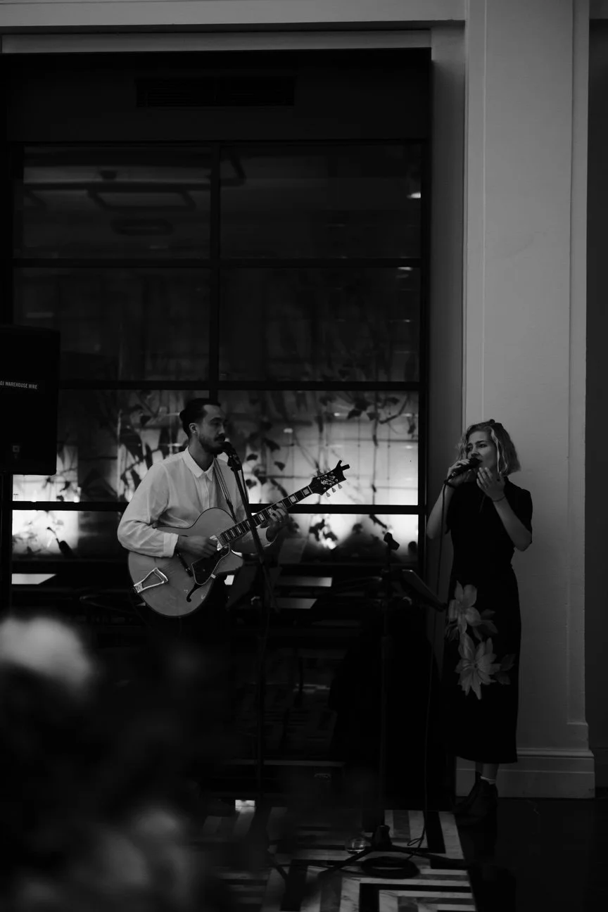 A black and white photo of Jess and Nick performing indoors, Nick playing an acoustic guitar and Jess singing into a microphone, standing near a large window.