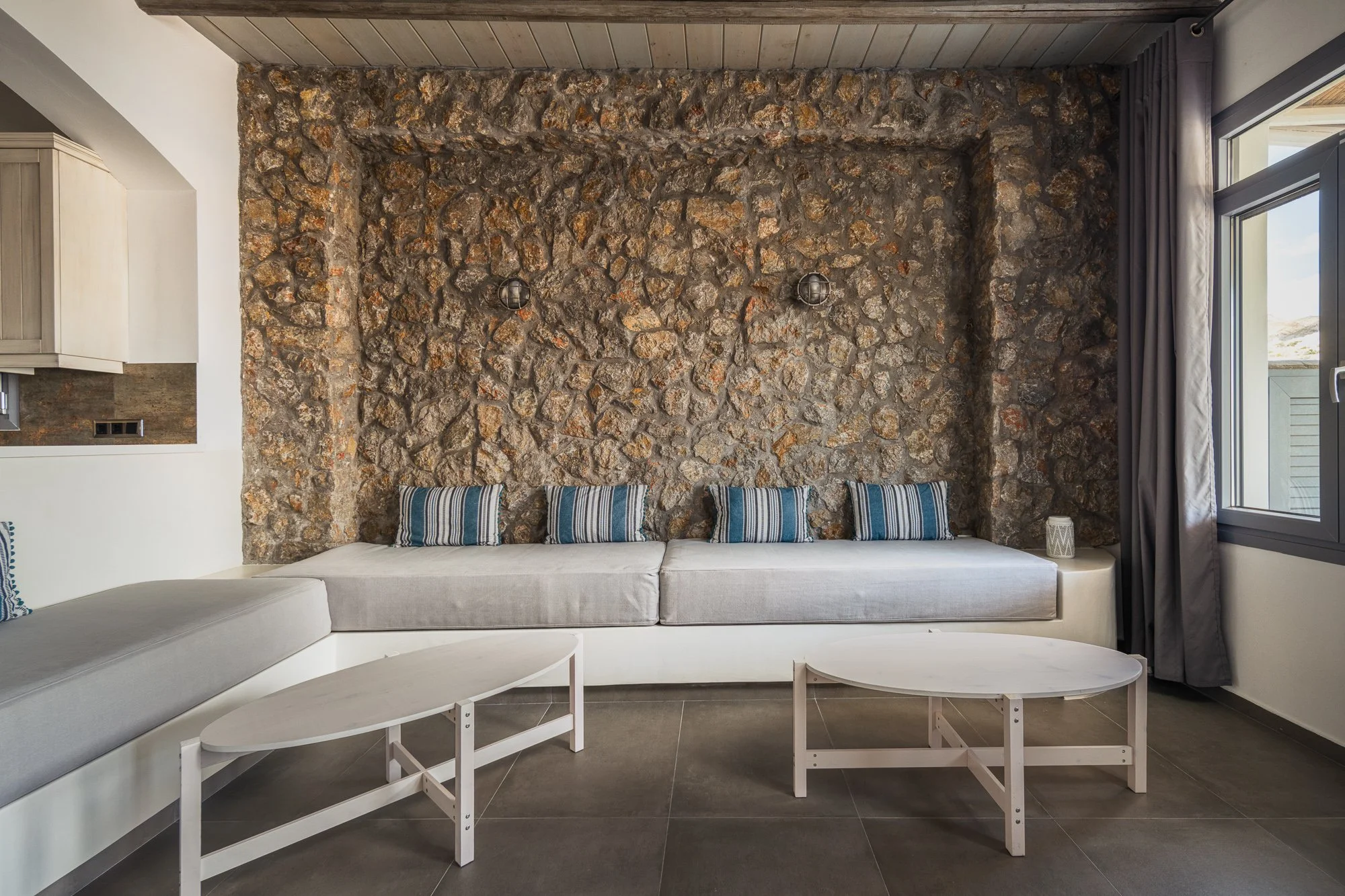 Living room with a stone accent wall, a white sectional sofa with striped pillows, two white coffee tables, and a window with gray curtains.