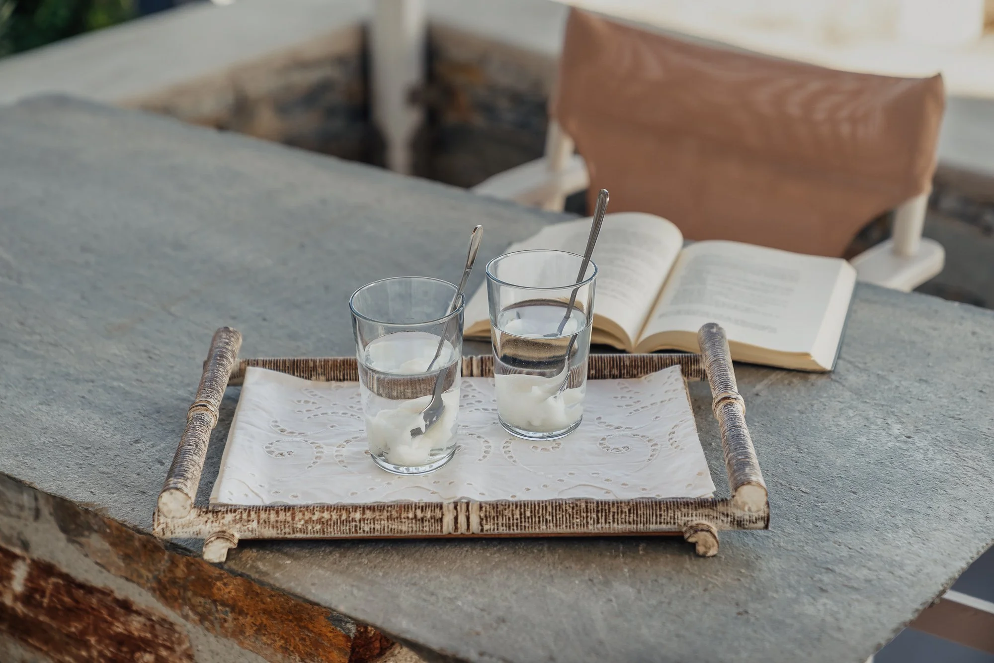 A tray holding two glasses of water with ice and spoons, placed on a table with a lace cloth, with an open book in the background.