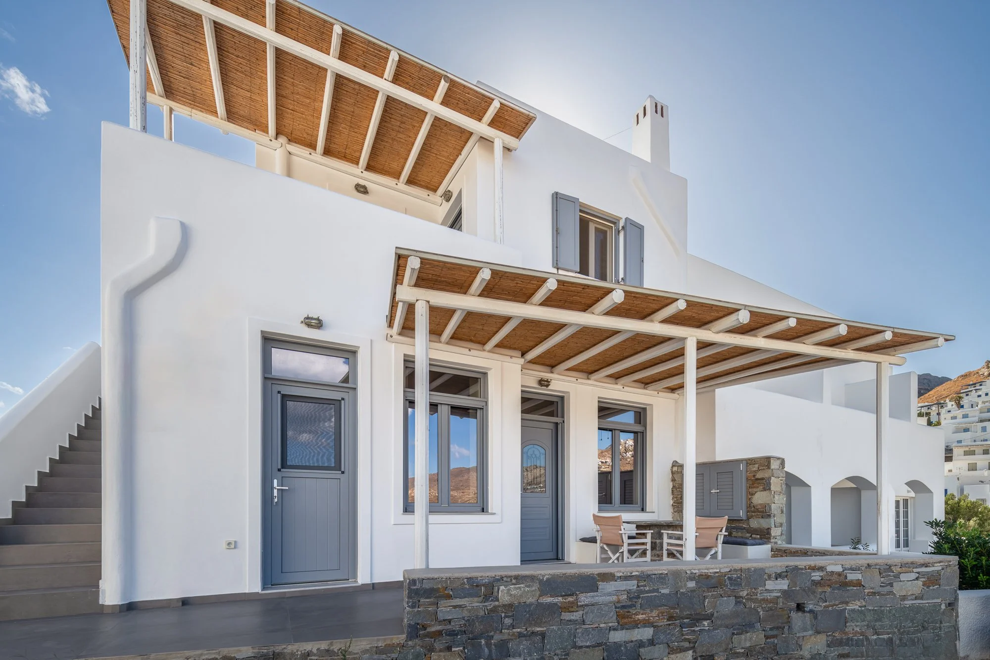 A white Mediterranean-style house with blue doors and windows, stone accents, a balcony with a wooden pergola, and an outdoor seating area under a wooden awning.