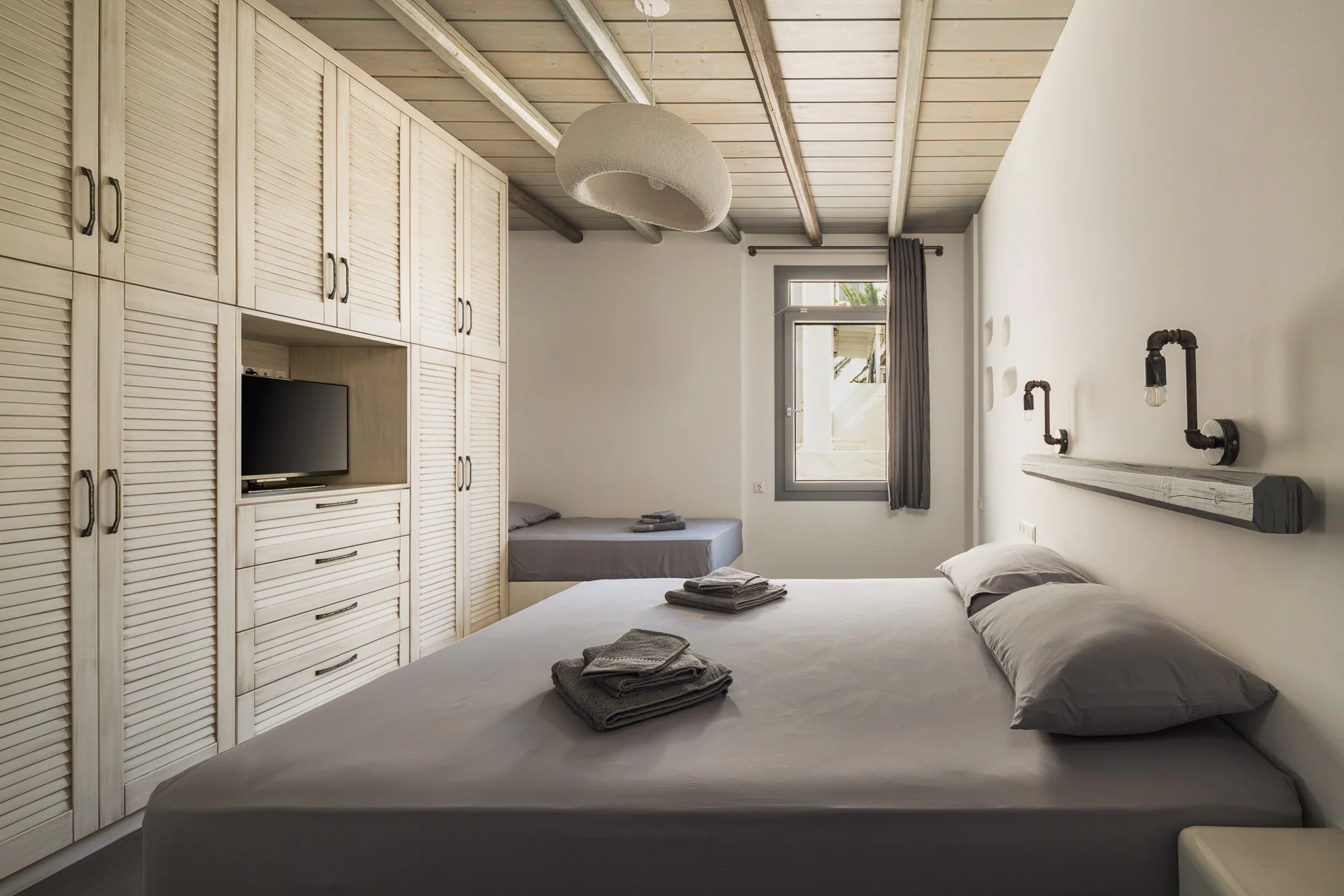 A bedroom with a large bed, two pillows, and folded towels on top, a smaller bed in the corner, wooden wardrobe with a TV, window with curtains, wall-mounted lamps with exposed bulbs, and a wooden plank on the wall.