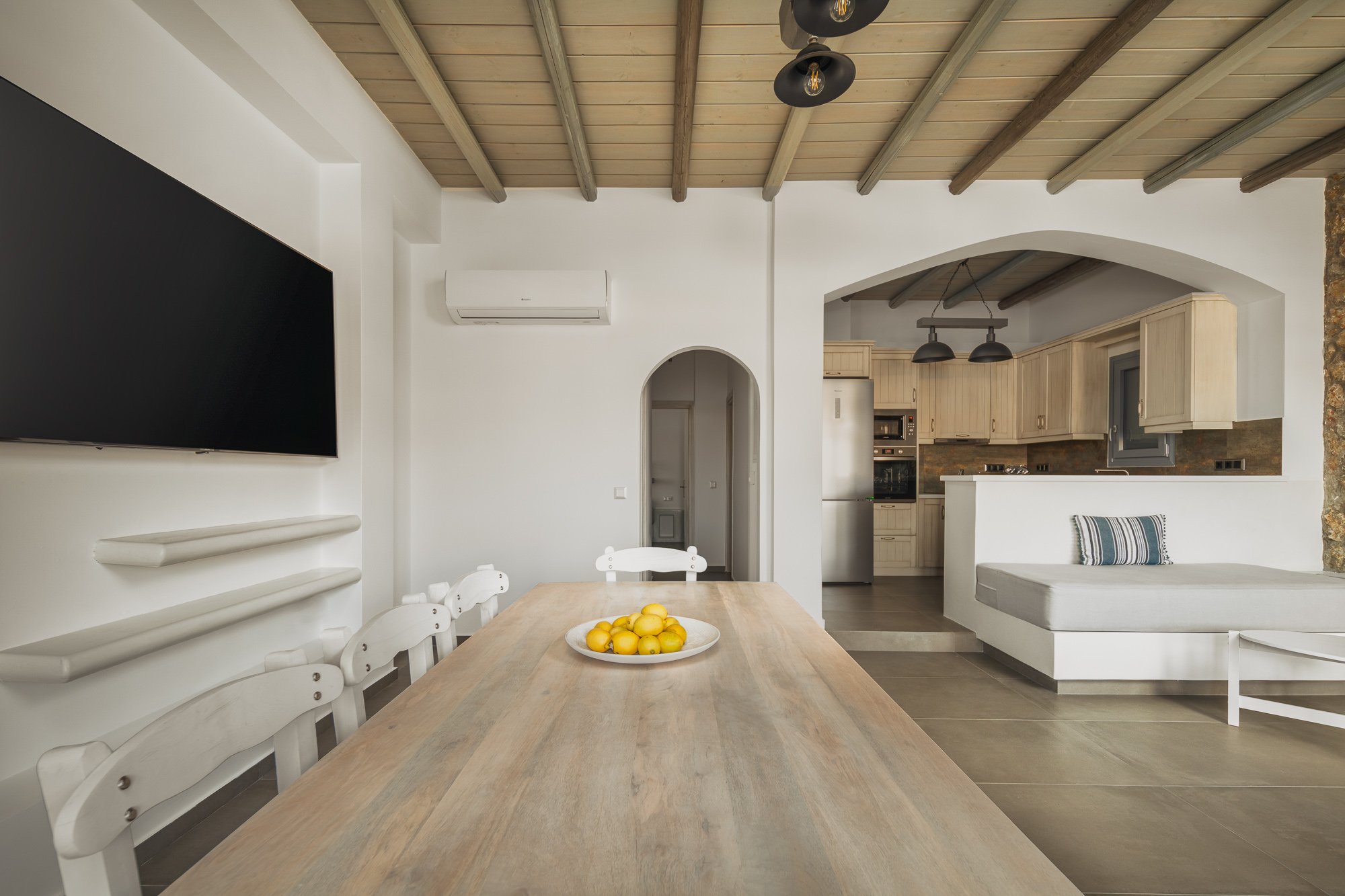 Dining area with a wooden table and white chairs, a bowl of lemons, a large flat-screen TV on the wall, and a kitchen in the background with light cabinetry, a refrigerator, and black pendant lights