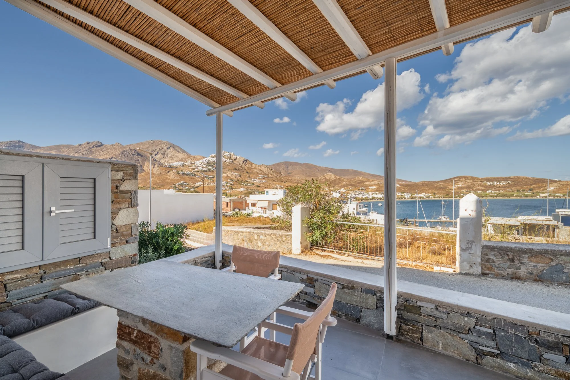 A covered outdoor patio with a stone table, surrounded by chairs, overlooking a coastal landscape with hills, houses, and boats on the water, under a partly cloudy sky.