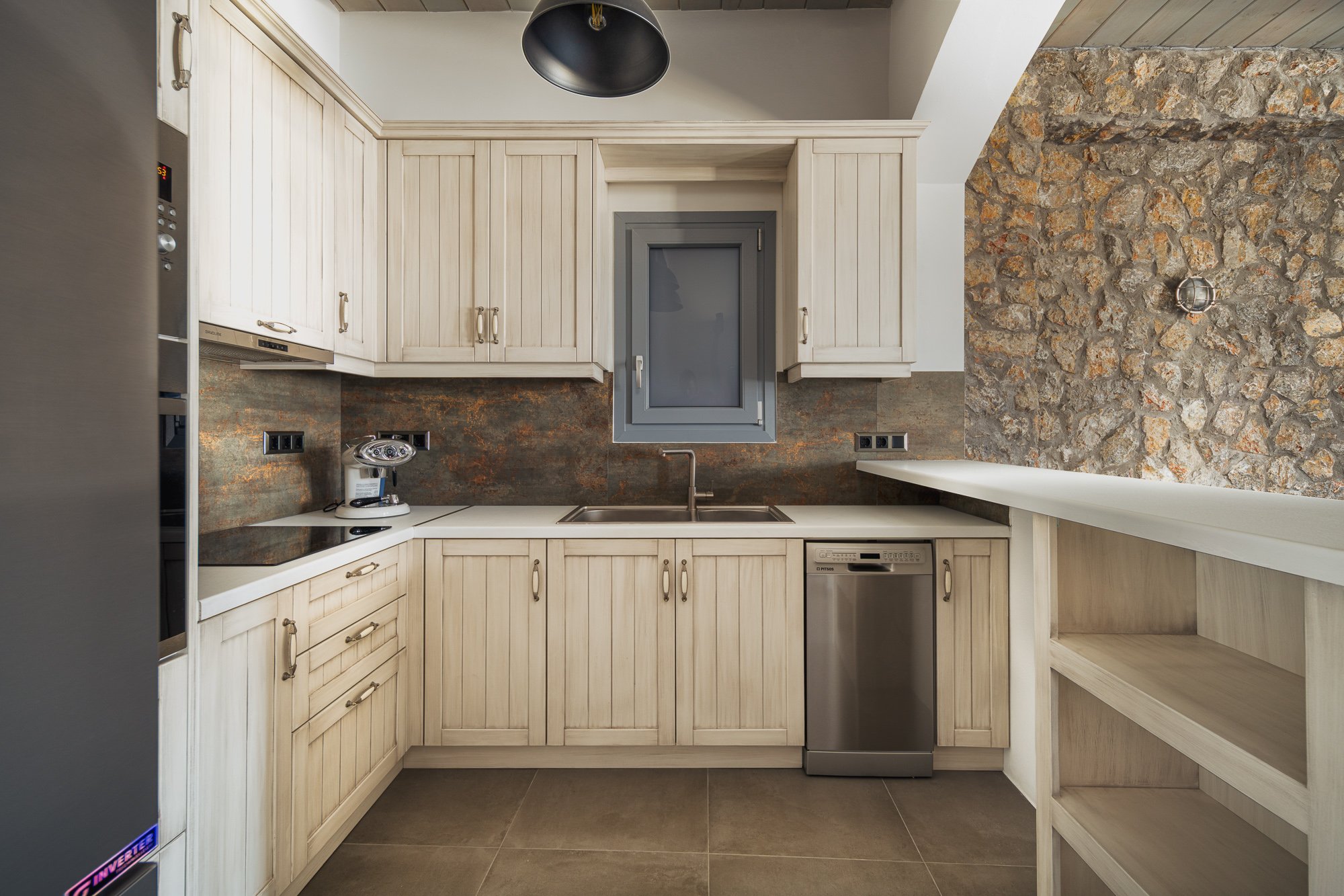 Small kitchen with beige cabinetry, a gray window, dark backsplash, a coffee maker, and a dishwasher, with a stone wall on the right and gray tile flooring.