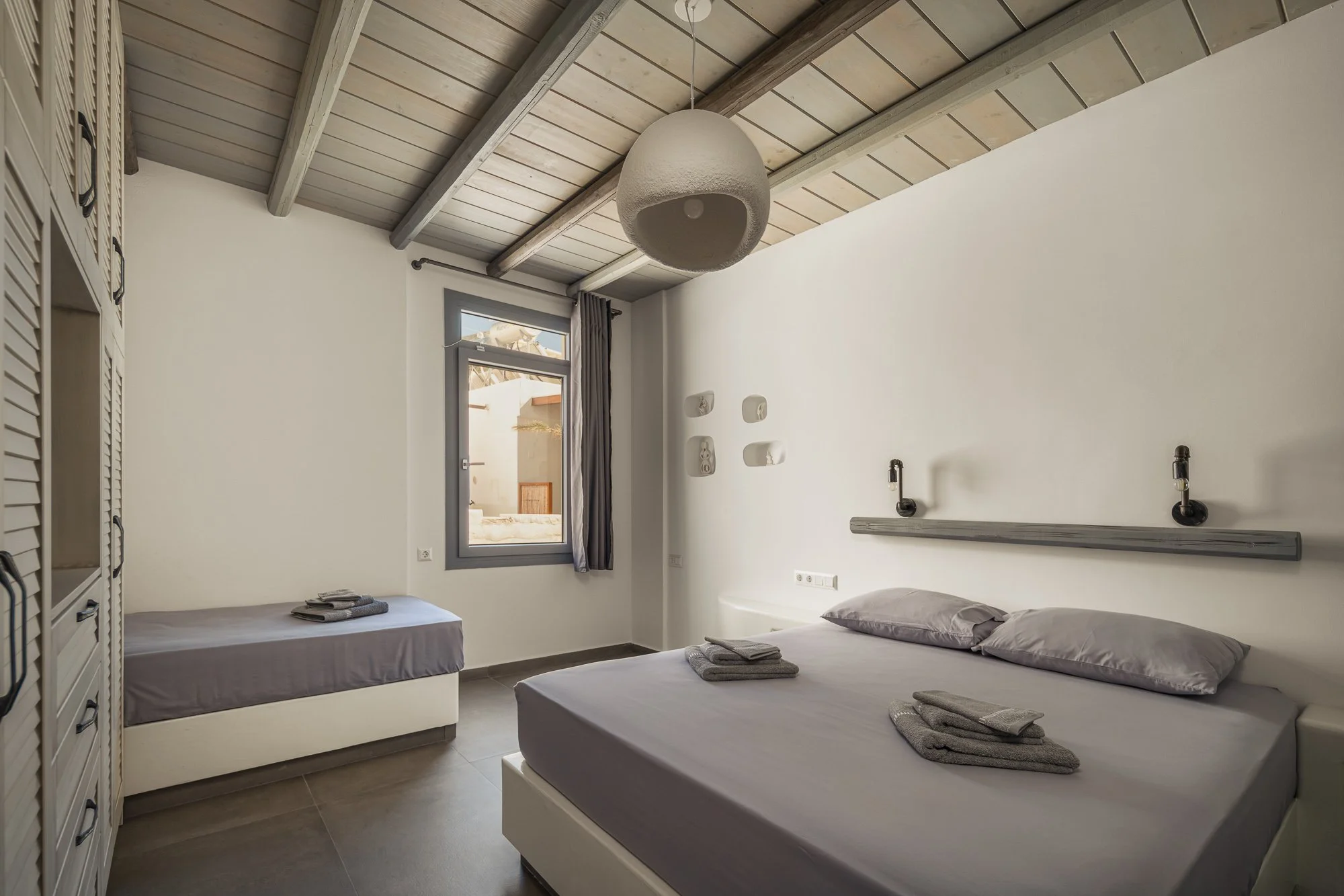 A bedroom with a sloped wooden ceiling, white walls, two beds with gray linens and towels, a window with gray trim and curtains, and modern decor elements.
