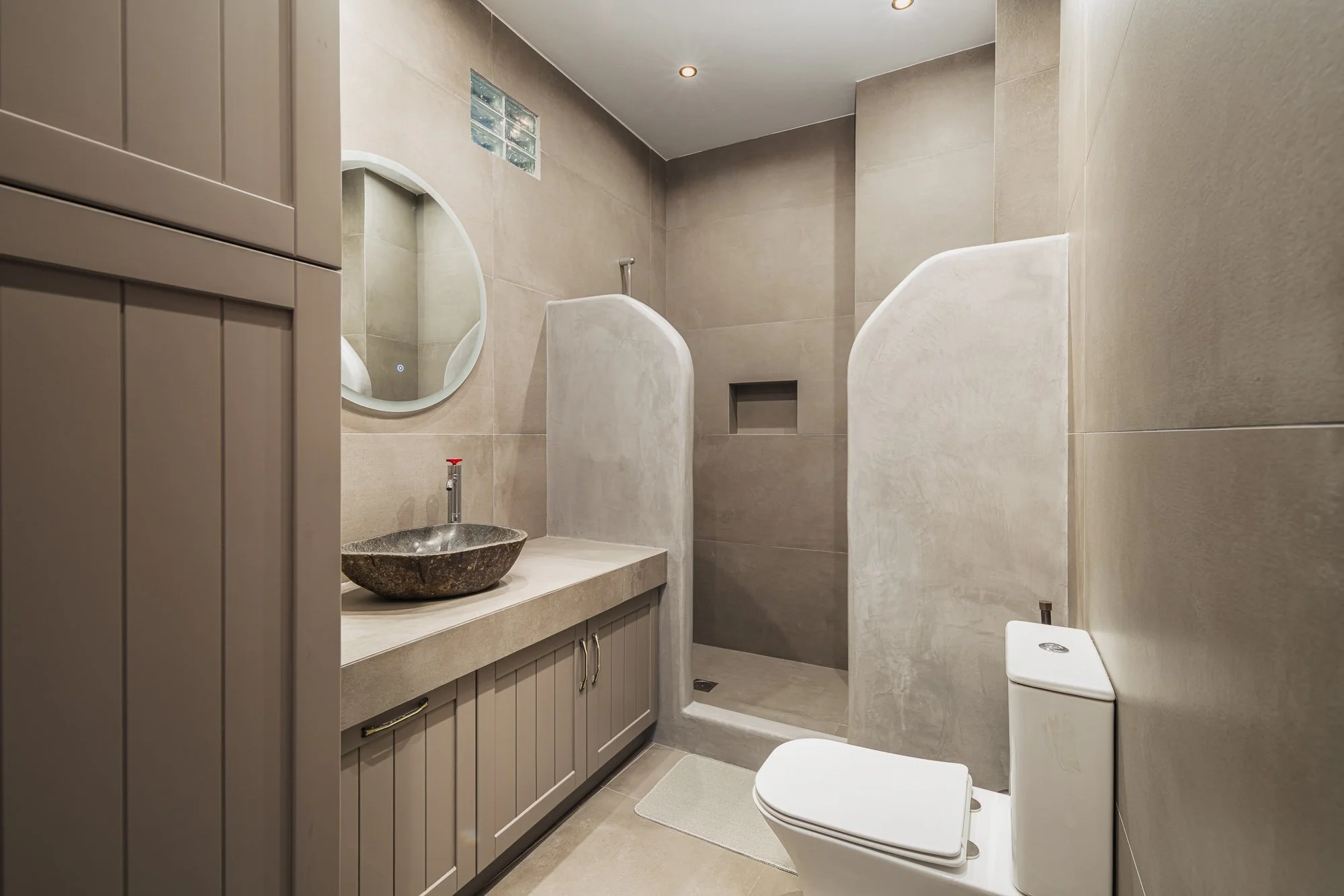 Modern bathroom with beige tiles, a gray vanity with a stone vessel sink, a round mirror, a walk-in shower with a partial wall, and a toilet.