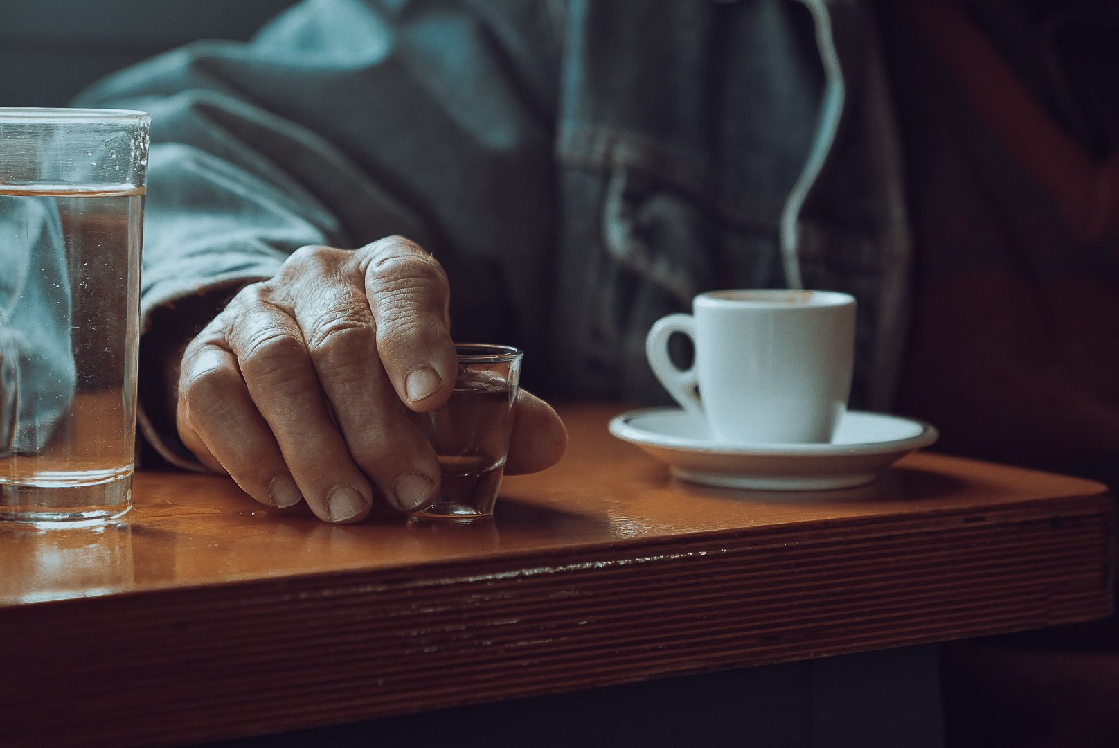 A person with weathered hands stretching over a wooden table, holding a small glass of liquor. There is a glass of water and a white cup of coffee or tea on a matching saucer on the table.