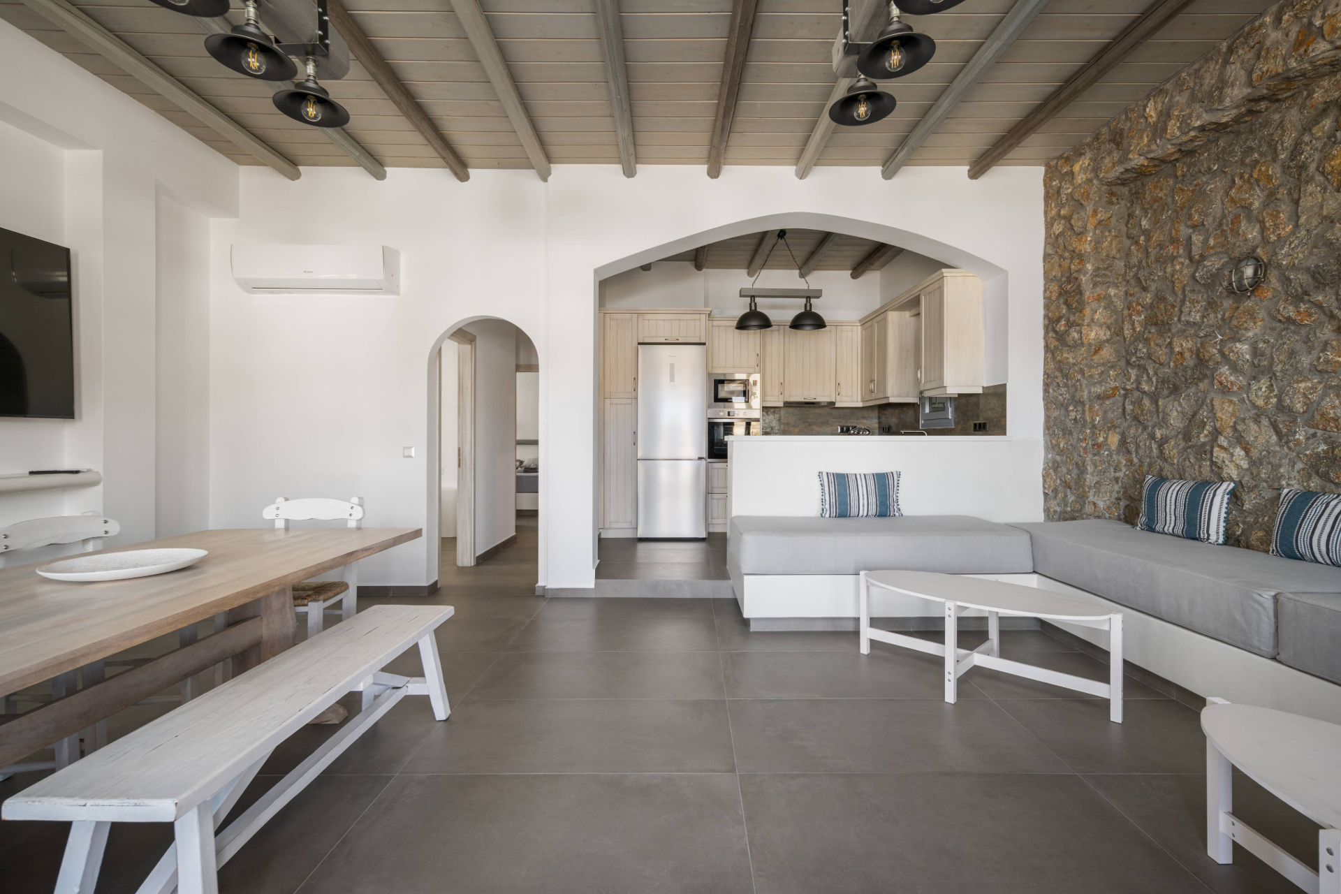 Open-concept living room and kitchen with white walls, stone accent wall, gray tiled floor, and wooden ceiling with black lighting fixtures. The kitchen has white cabinetry, stainless steel appliances, and a pass-through window. The living room contains a built-in gray sectional sofa with striped pillows, a white coffee table, and a dining area with a wooden table, benches, and chairs.