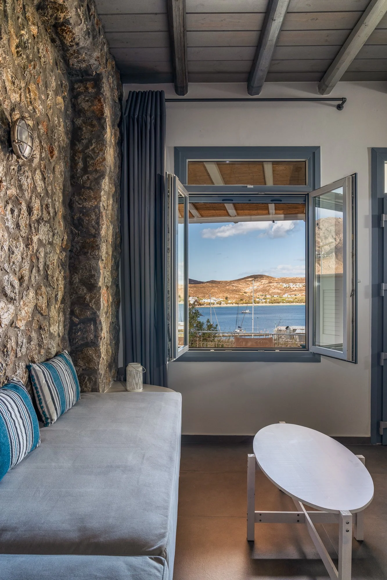 Interior of a room with stone wall, open window overlooking a body of water with sailboats and hills in the distance, white sofa with blue striped pillows, small white table.