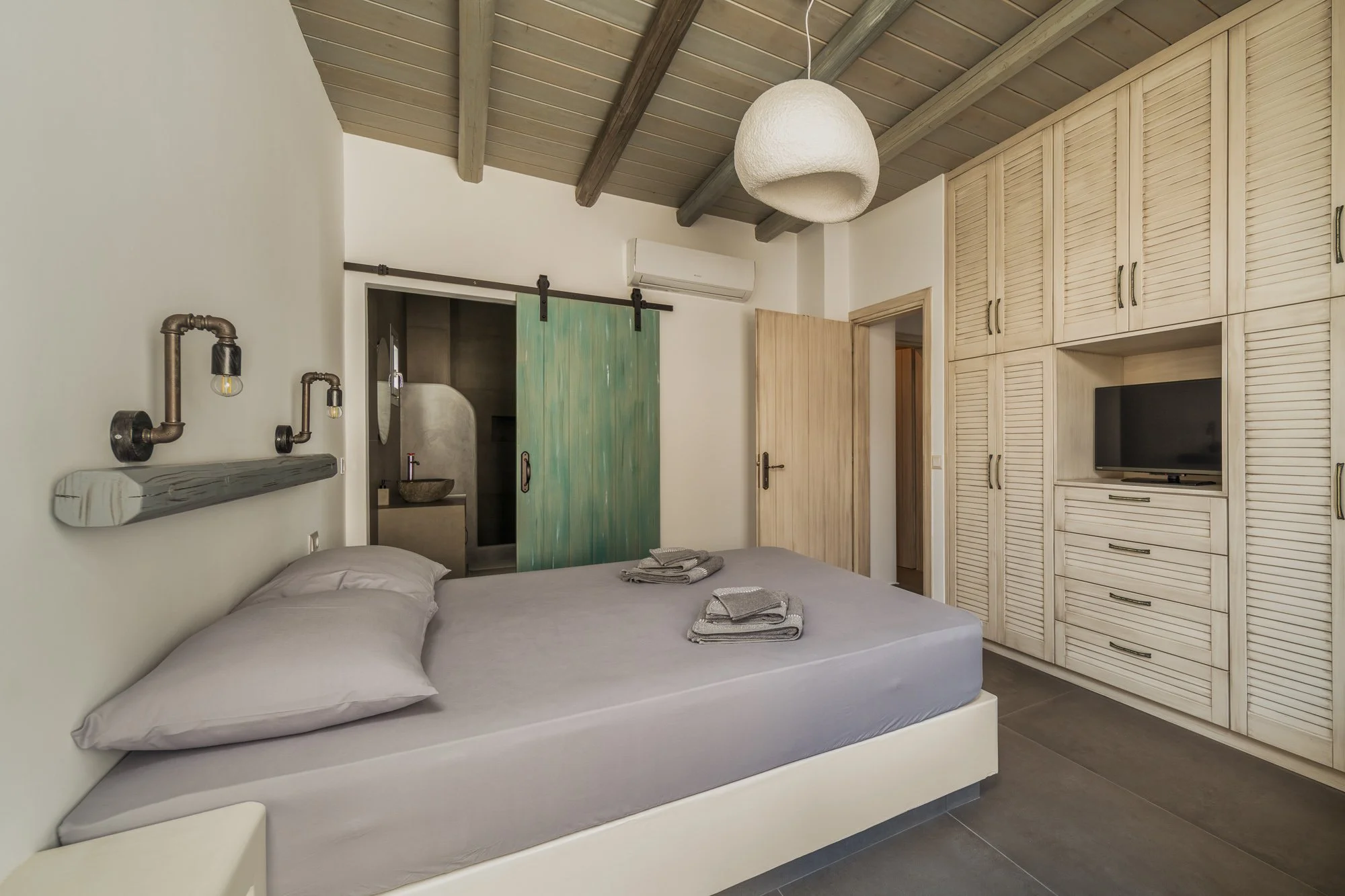 Modern bedroom with a bed, gray sheets, and pillows, featuring unique lighting fixtures on the wall, a wooden ceiling with beams, a sliding green door leading to a small bathroom, built-in white wooden cabinets with a TV, and a wooden door.