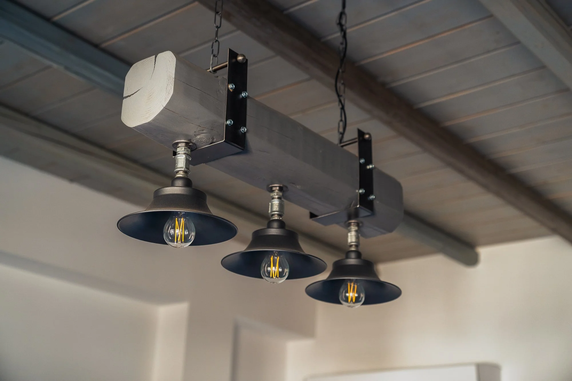A rustic pendant light fixture with three black metal shades and exposed filament light bulbs, mounted on a wooden ceiling with visible beams.