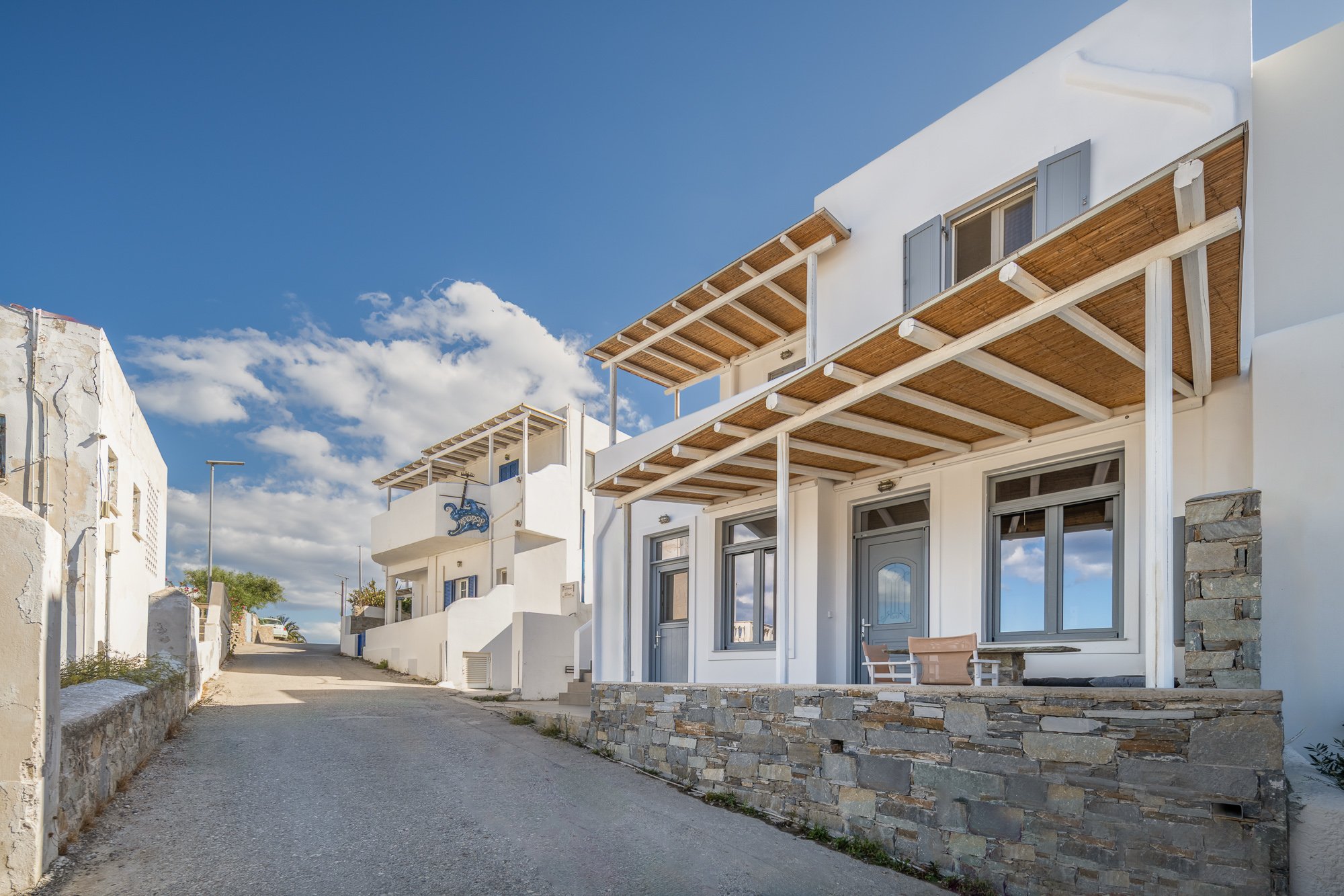 White Mediterranean-style houses with stone and wood accents, small balconies, and a dirt road under a partly cloudy blue sky.