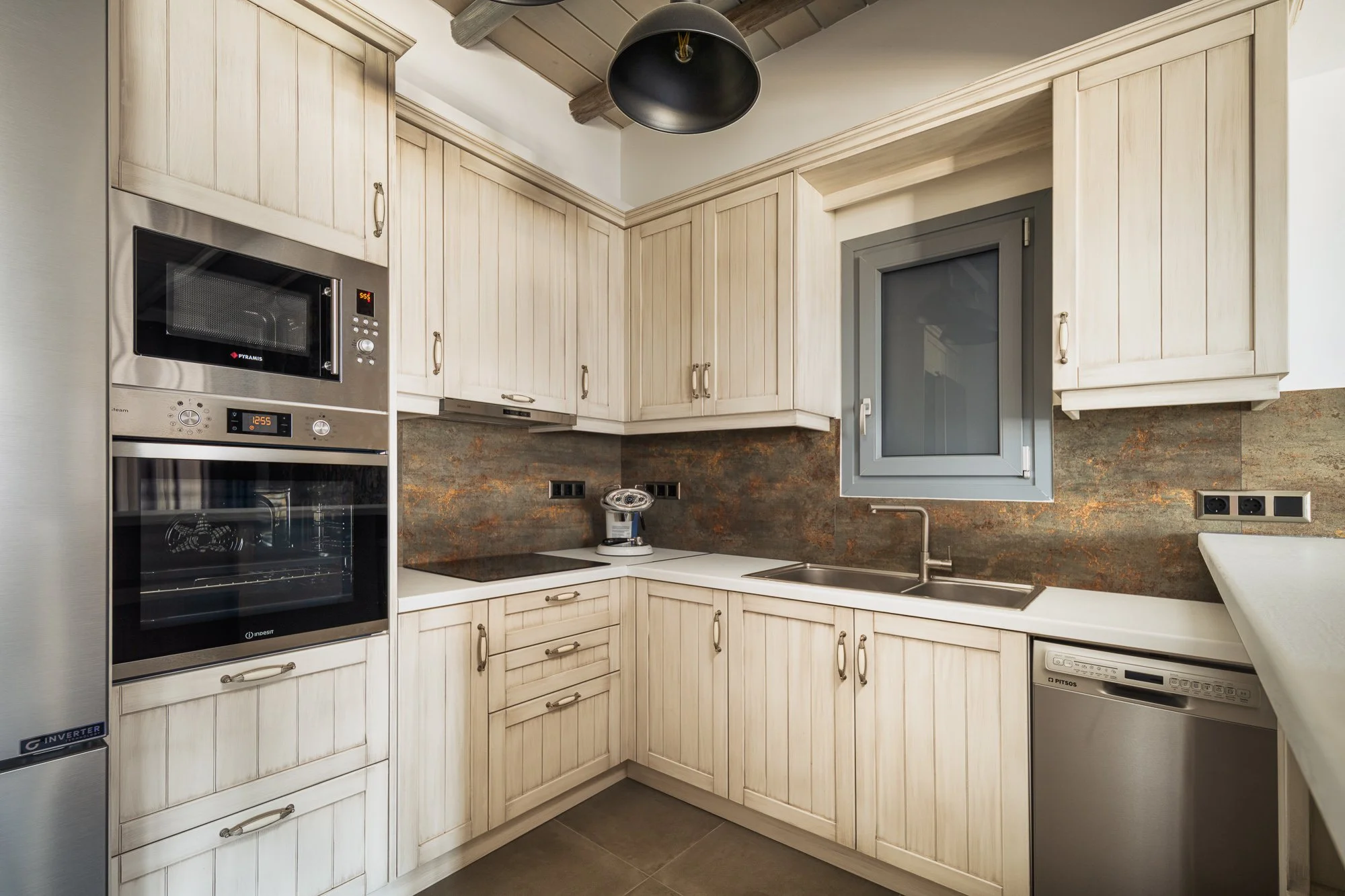 Kitchen with light wood cabinets, black pendant light, stainless steel appliances, gray window, and rust-colored backsplash.
