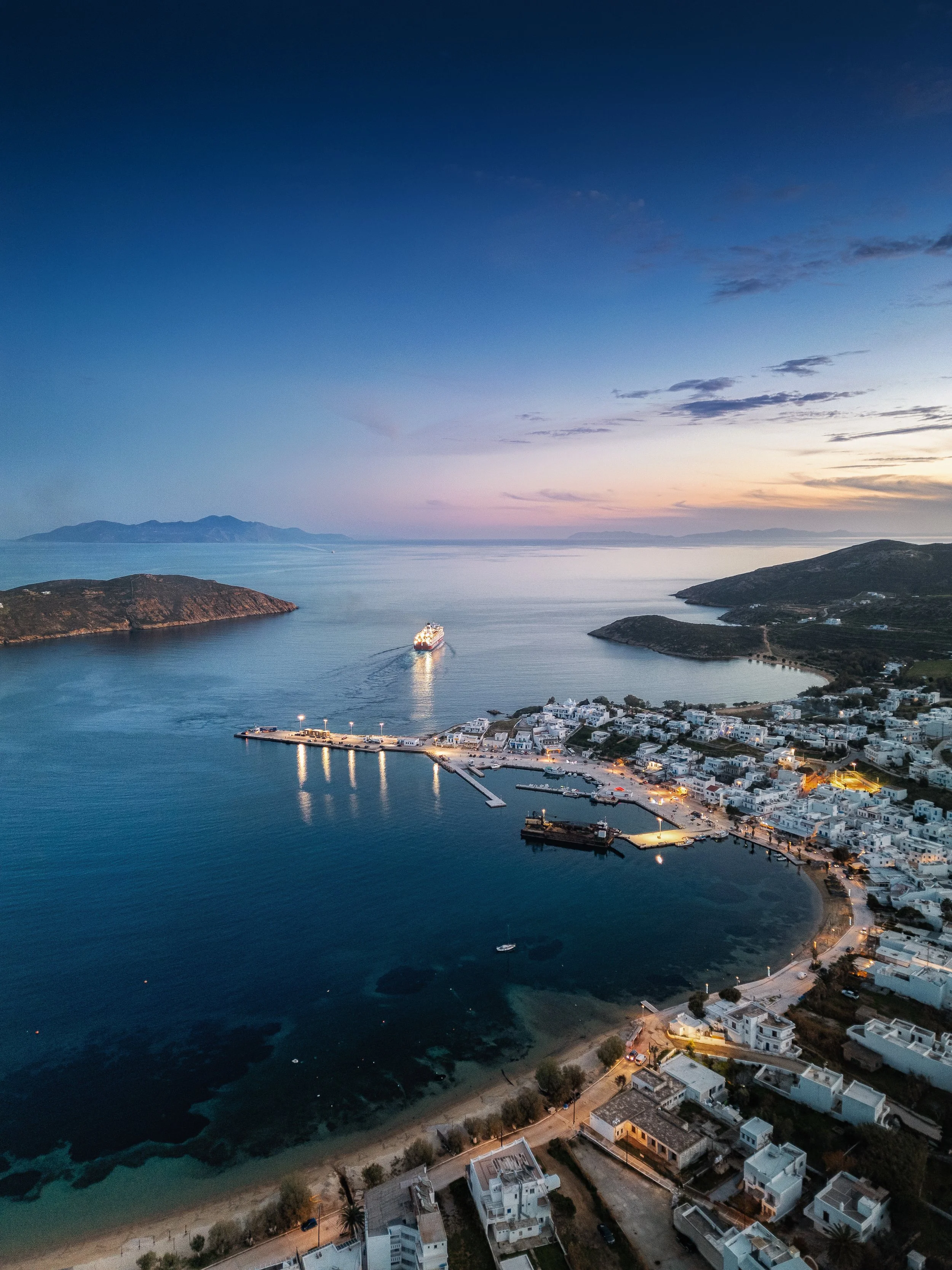 Aerial view of a coastal town at dusk with a harbor, white buildings, and a cruise ship sailing in the water.