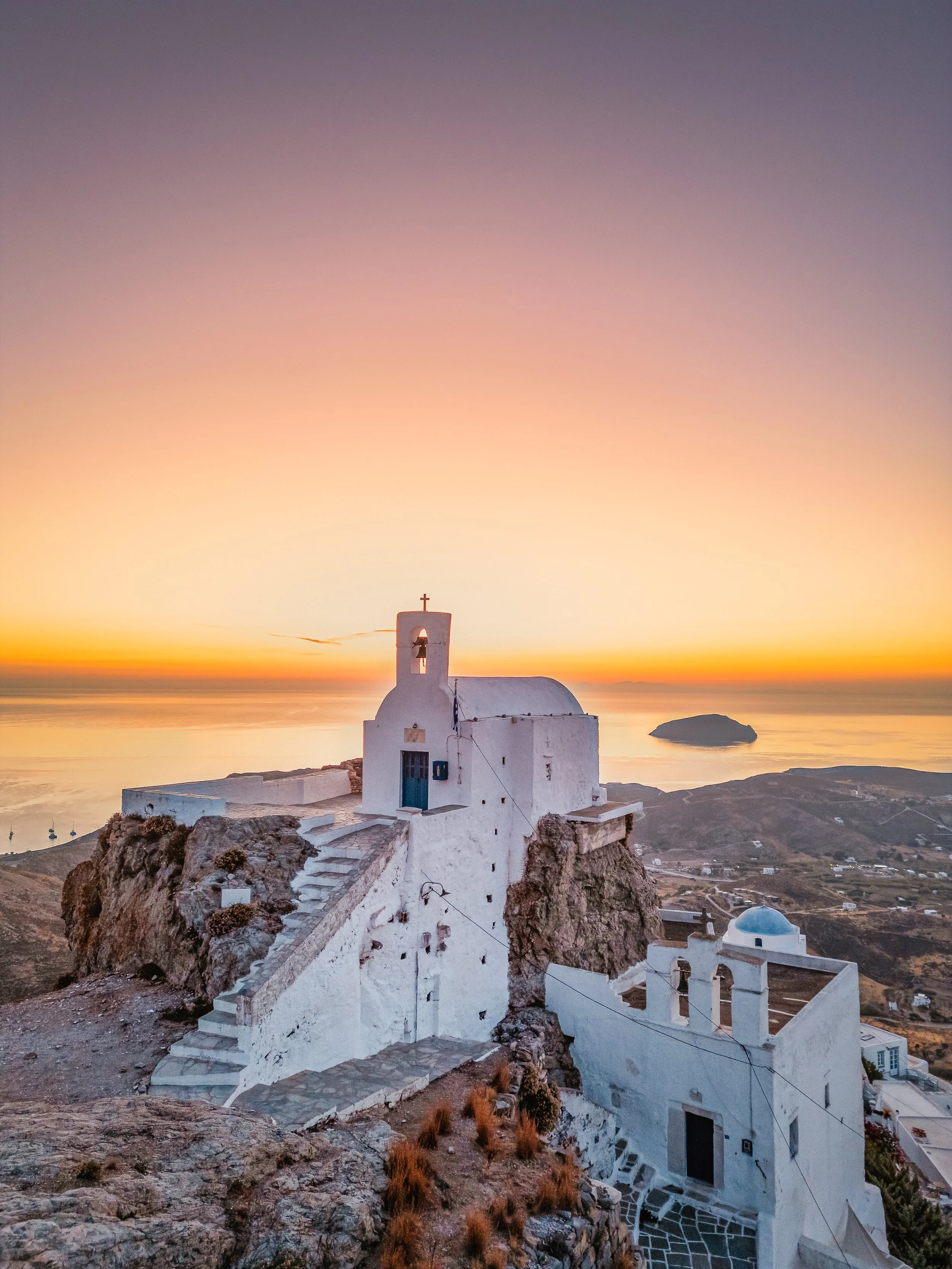 A white chapel built into a rocky hillside during sunset, overlooking the ocean with a small island in the distance.