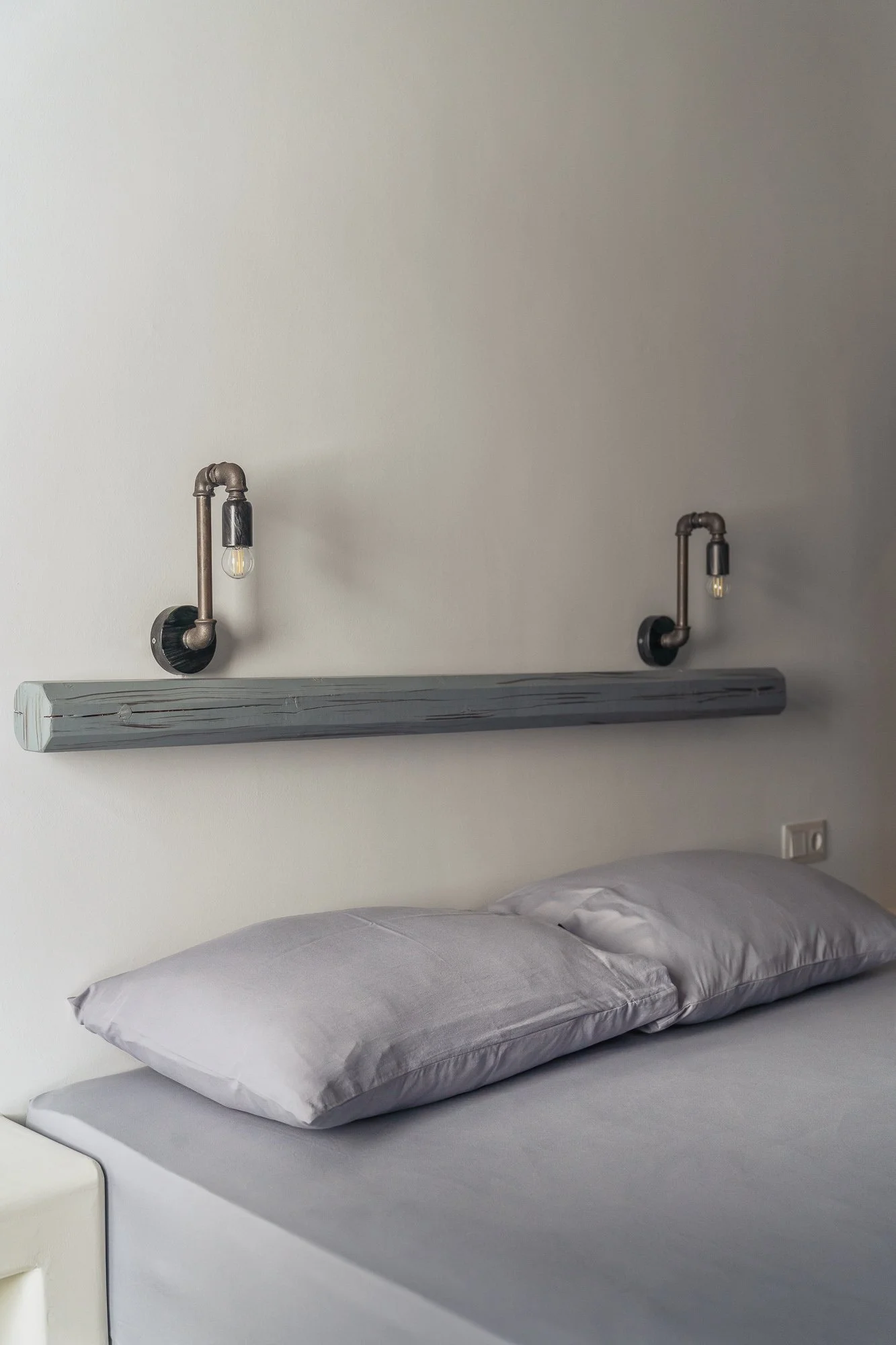 Bed with gray pillows and bedsheet, wall-mounted lamp fixtures made of pipes with light bulbs above the headboard, and electrical outlet on the wall.