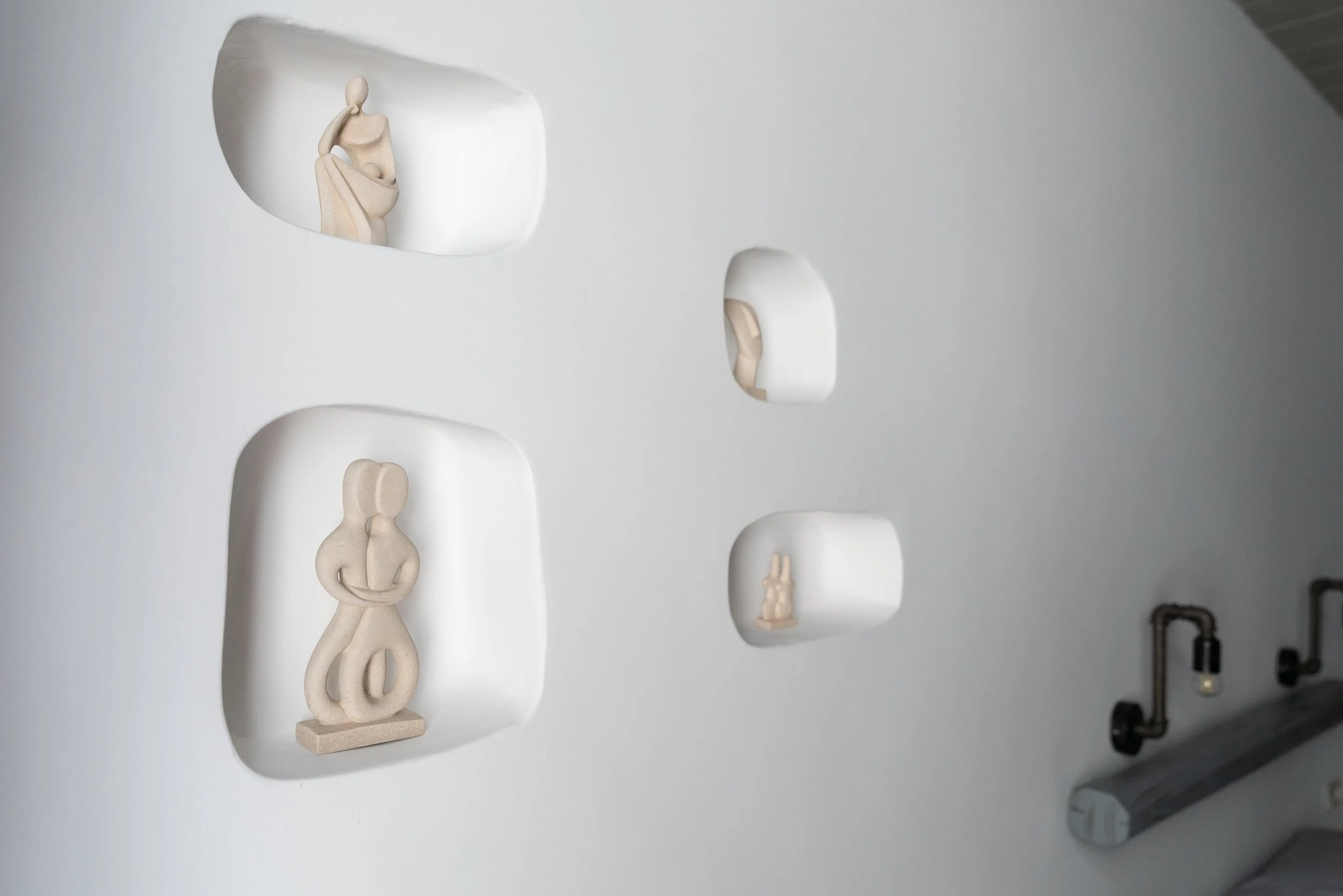 White wall with four embedded display niches holding abstract beige sculptures of human figures and forms, with a part of a metallic pipe on the lower right.