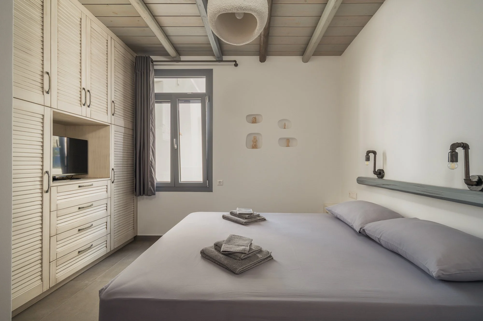 A bedroom with a bed, folded towels on the bed, a window, built-in wardrobe, and minimalist decor.