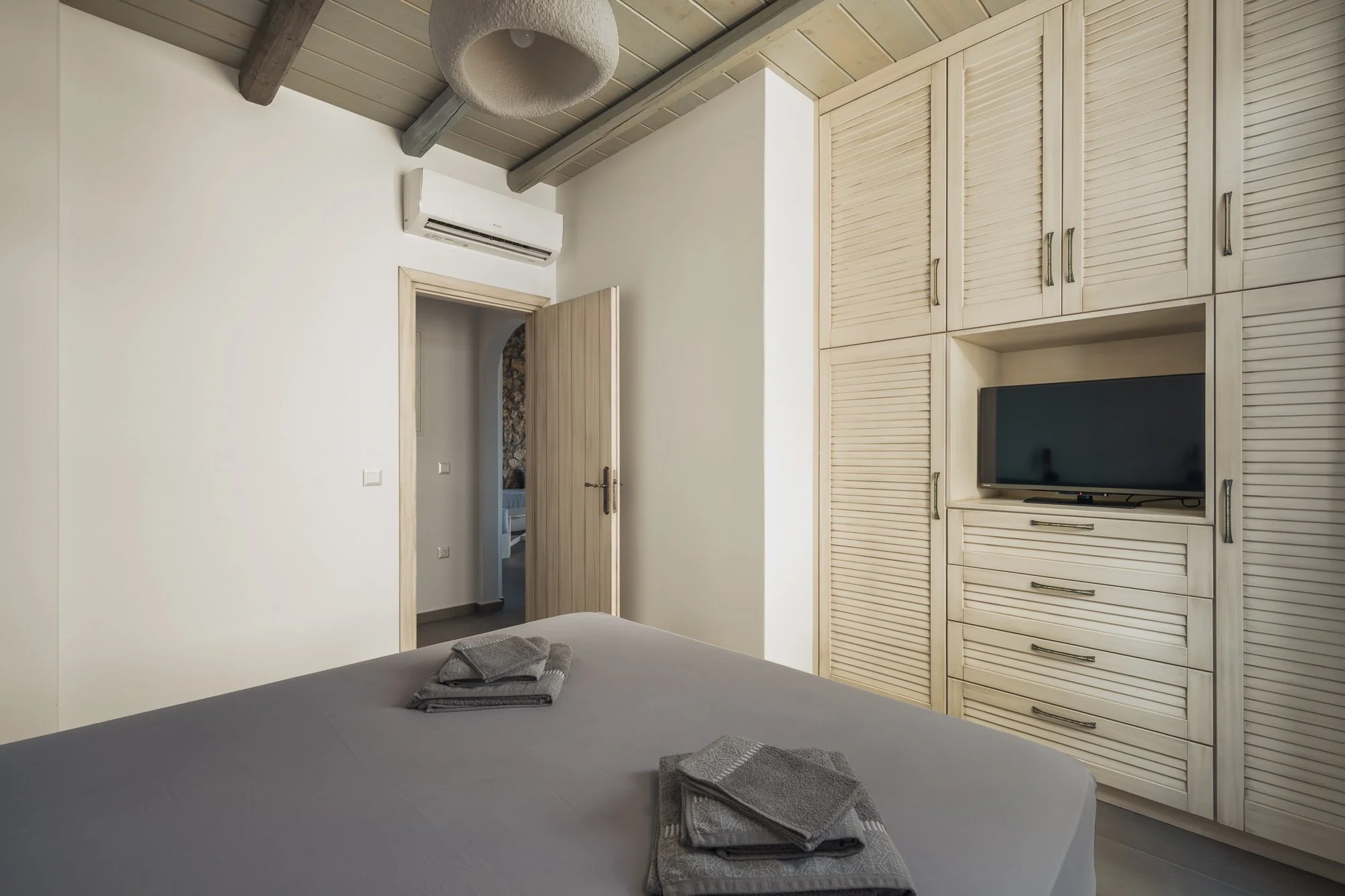 A bedroom with a bed, towels on the bed, a large wooden wardrobe, a small television, an air conditioning unit, and a door leading to another room.