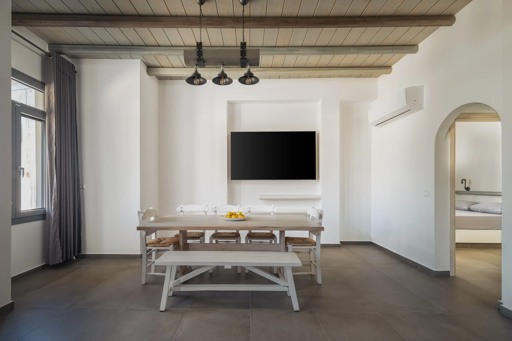 Modern dining room with a rectangular wooden table, white chairs with woven seats, and a bench. A bowl of lemons is on the table. A large flat-screen TV is mounted on the wall. There is a window with curtains on the left, an air conditioning unit on the right, and a doorway to a bedroom.