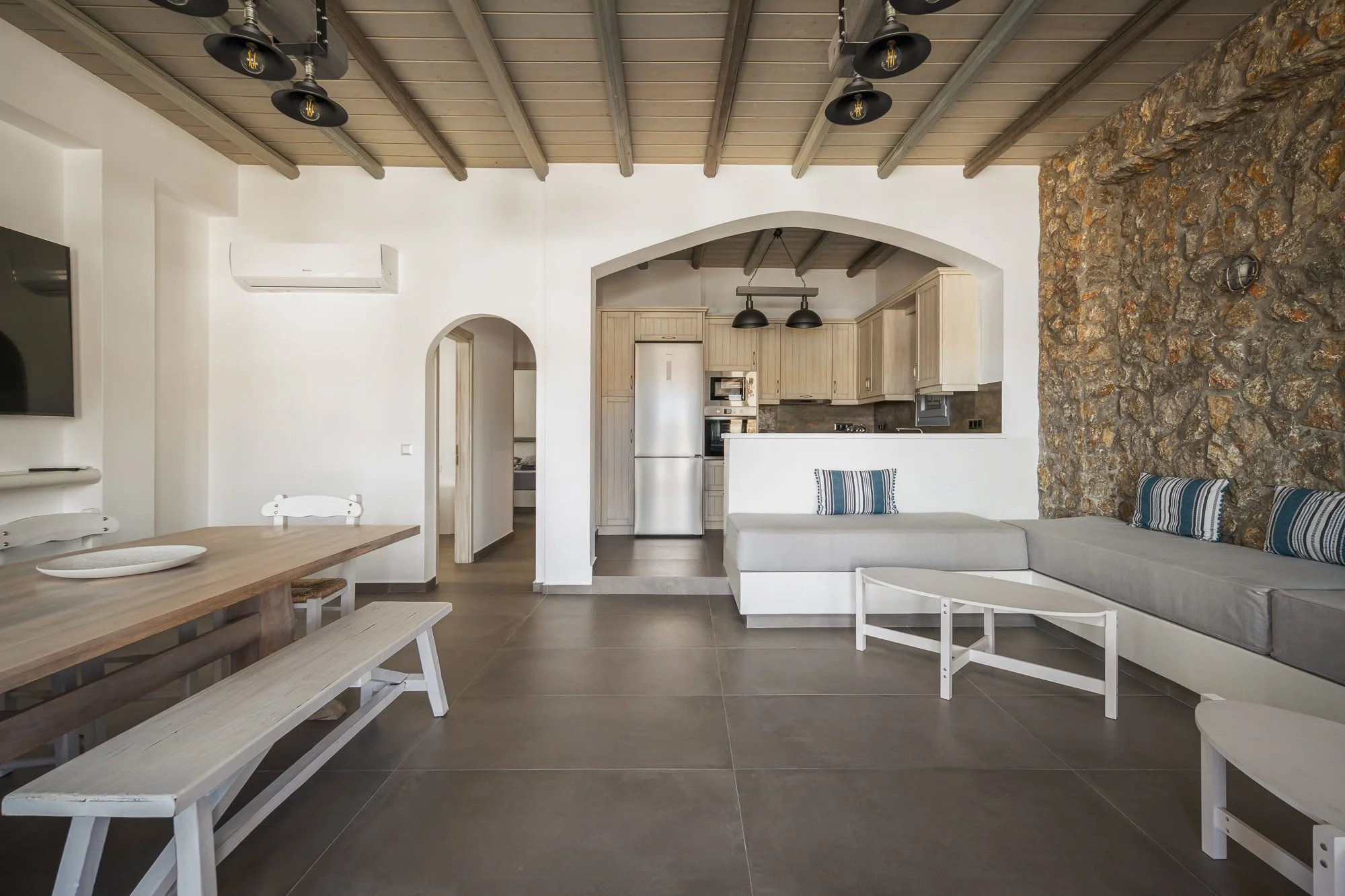Living room with stone accent wall, white furniture, wooden dining table with benches, and a kitchen with beige cabinets and stainless steel appliances in the background, under a wooden ceiling with black light fixtures.