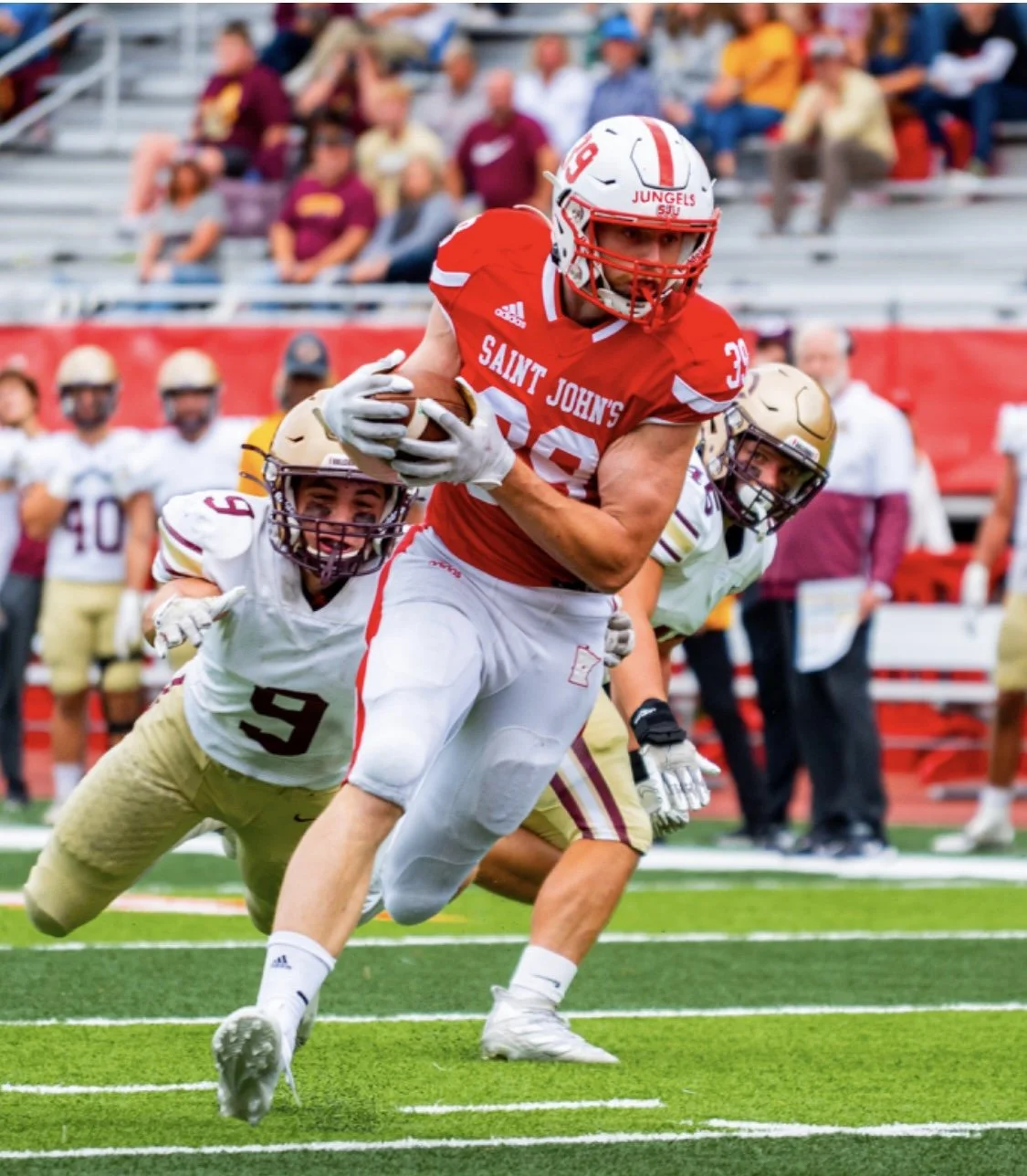 Football player in red jersey from Saint John's University running with the ball while being chased by defenders in white and gold jerseys during a game.