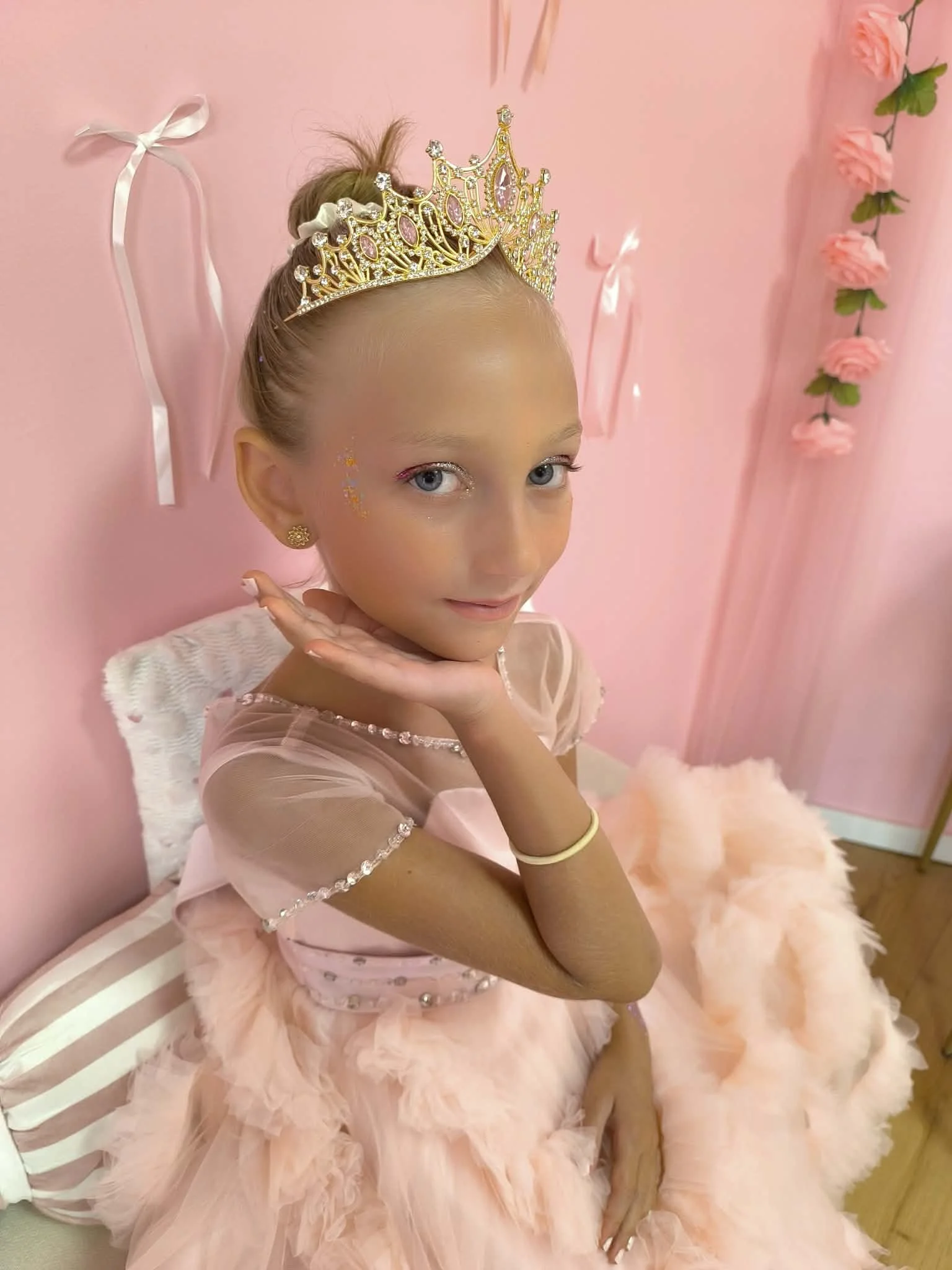 Une jeune fille habillée en tutu rose, portant une couronne dorée et des accessoires de fête, assise dans une pièce rose décorée avec des fleurs en papier et des rubans.