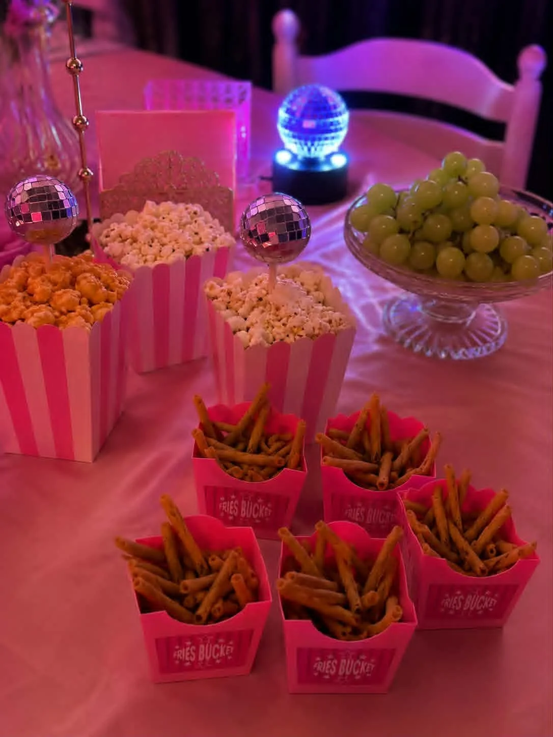 Table avec des cannes de popcorn dans des boîtes roses, des petits frites dans des boîtes roses, un grand bol de raisins verts, et une boule à facettes illuminée en arrière-plan.