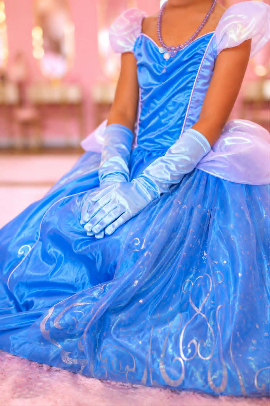 Fille en costume de princesse bleue avec des gants longs et un collier, assise sur un tapis rose dans une pièce décorée en rose.