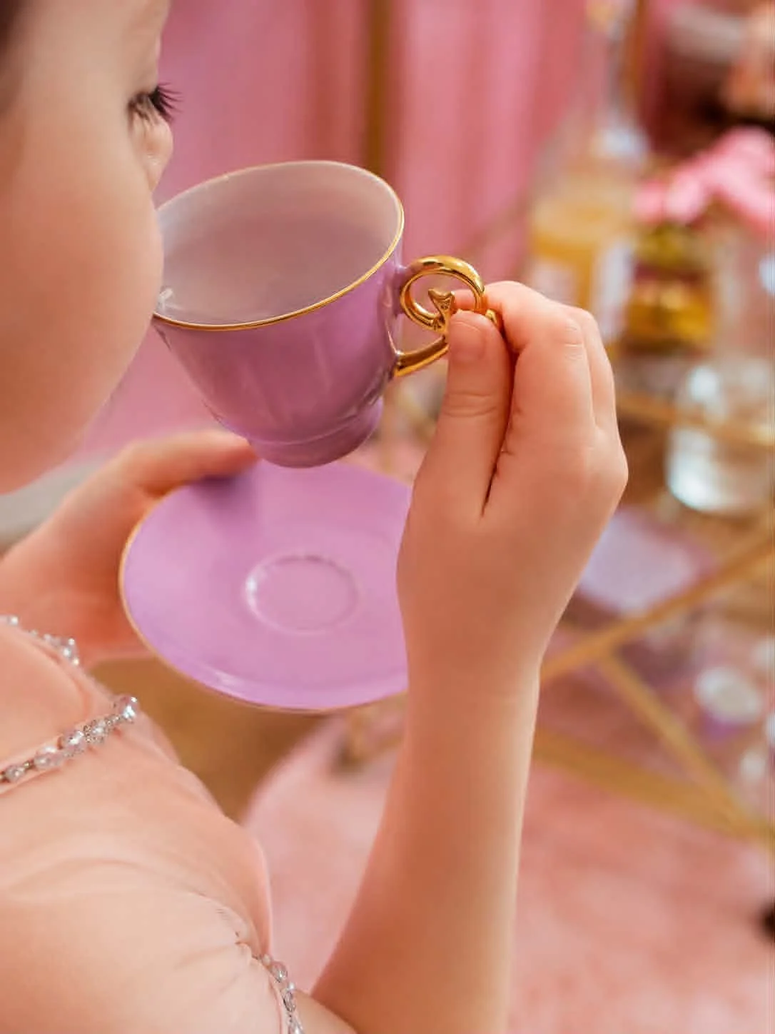 Enfant tenant une tasse rose avec une poignée dorée, dégustant une boisson, dans un décor pastel et décoratif.